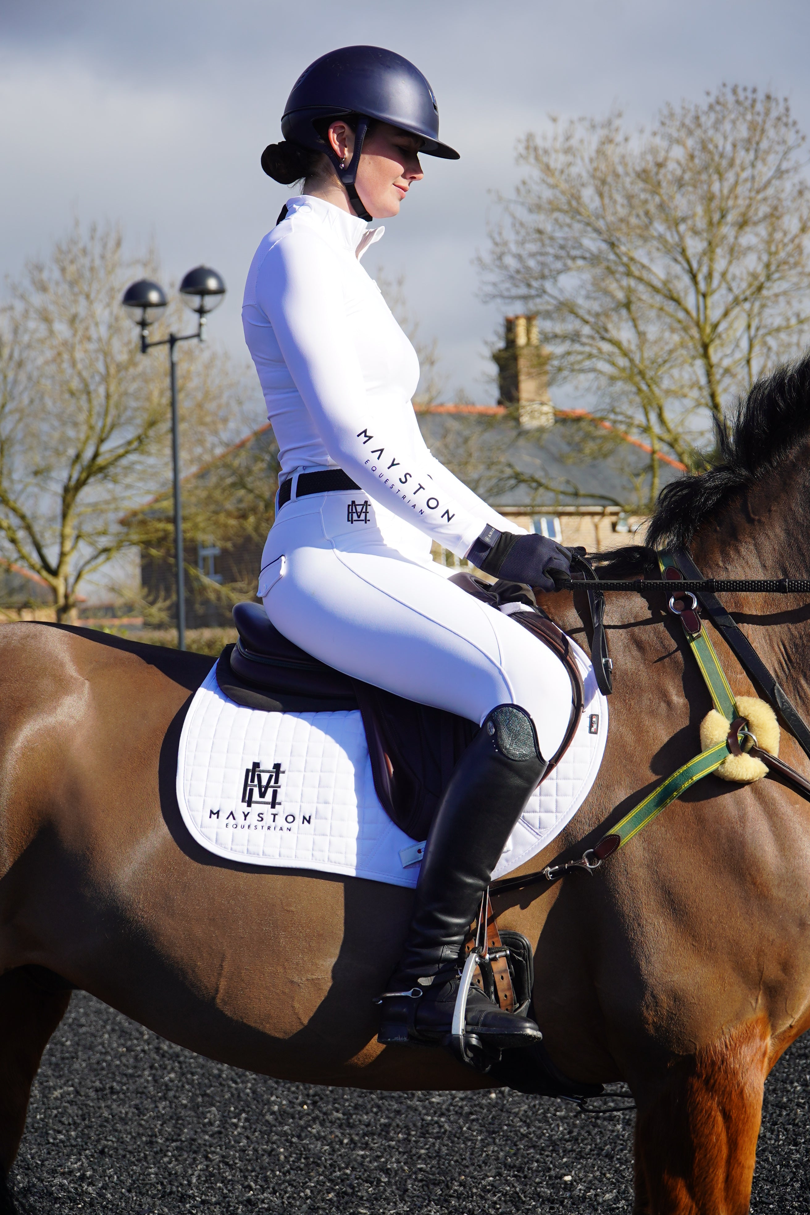 Person riding a horse with a blurred background. Sleek silhouette baselayer showshirt top subtle Mayston branding, maximum coverage without compromising style, next-level performance with a bold, minimalist look. Moisture-wicking, breathable fabric with four-way stretch, it keeps you cool and comfortable. Training, Everyday or competition wear. long sleeve Sleeve 1/4 zip with standup collar. Suble Black Mayston branding.