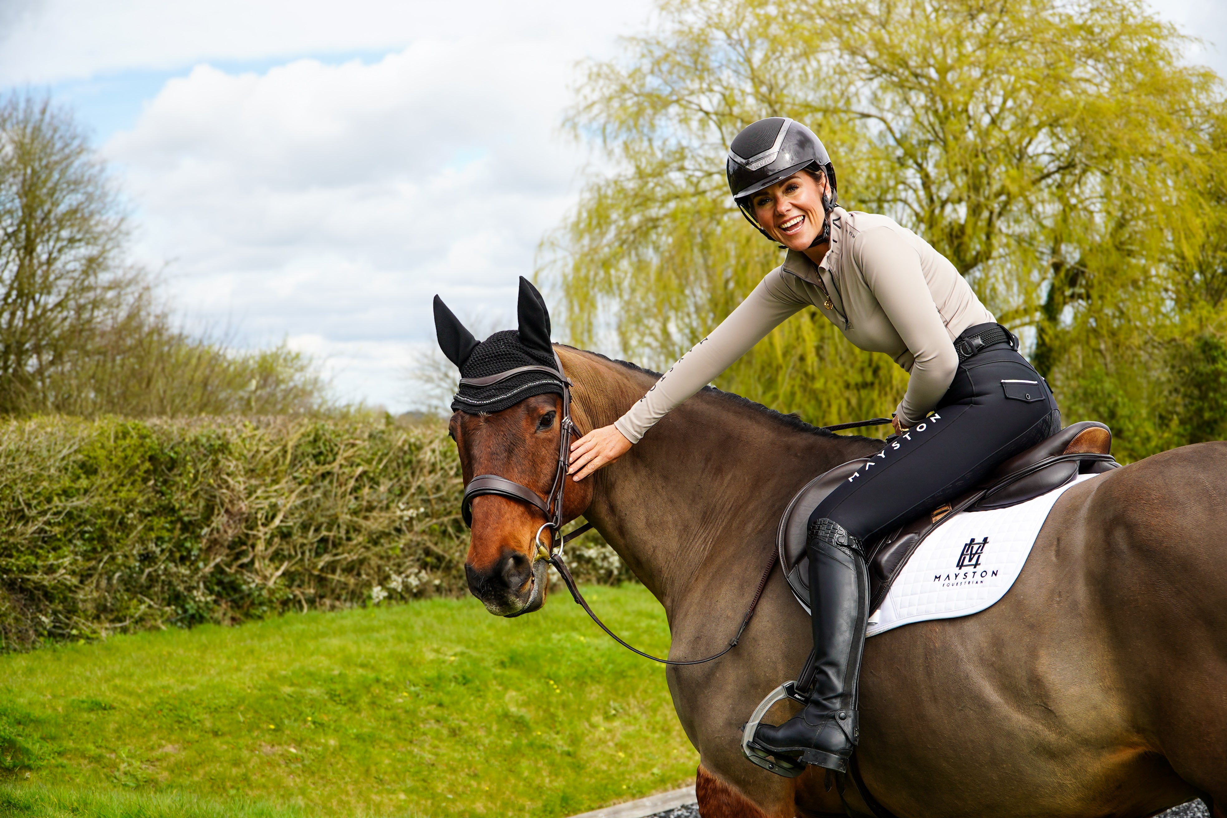 Woman riding a horse in an outdoor setting with trees and grass. classic equestrian fit with belt loops and secure fastening, combining traditional style with modern performance. Finished in sleek Classic Black with subtle white detailing and high-gloss silicone logos, they’re the perfect balance of elegance and functionality.