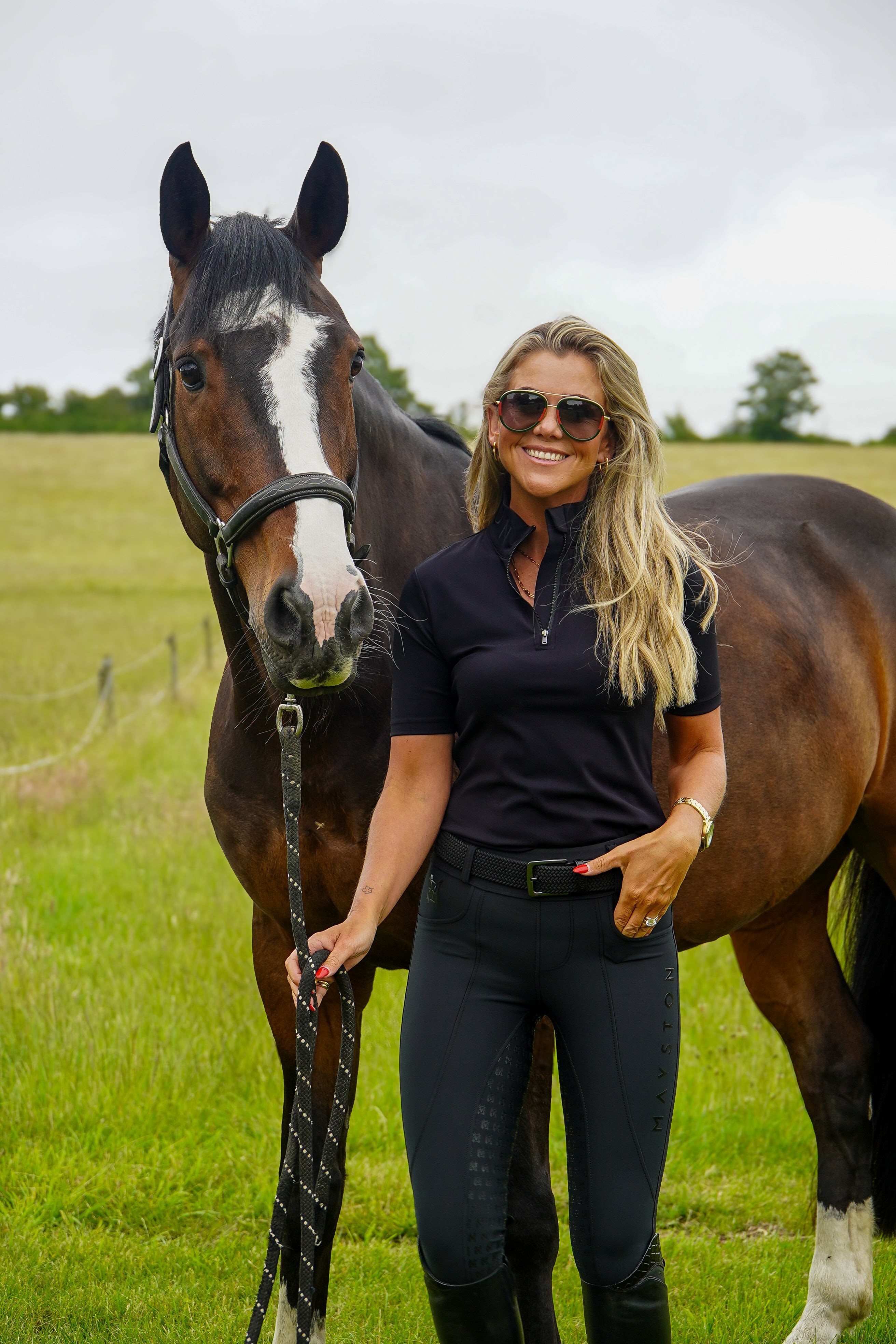 Woman in equestrian attire standing next to a horse in a field. Premium Black breeches with a Full Silicone seat. Stretch high waisted Jodhpurs. Deep pockets, supportive, comfortable and compression quality fabric. Elegant detailing with subtle branding.