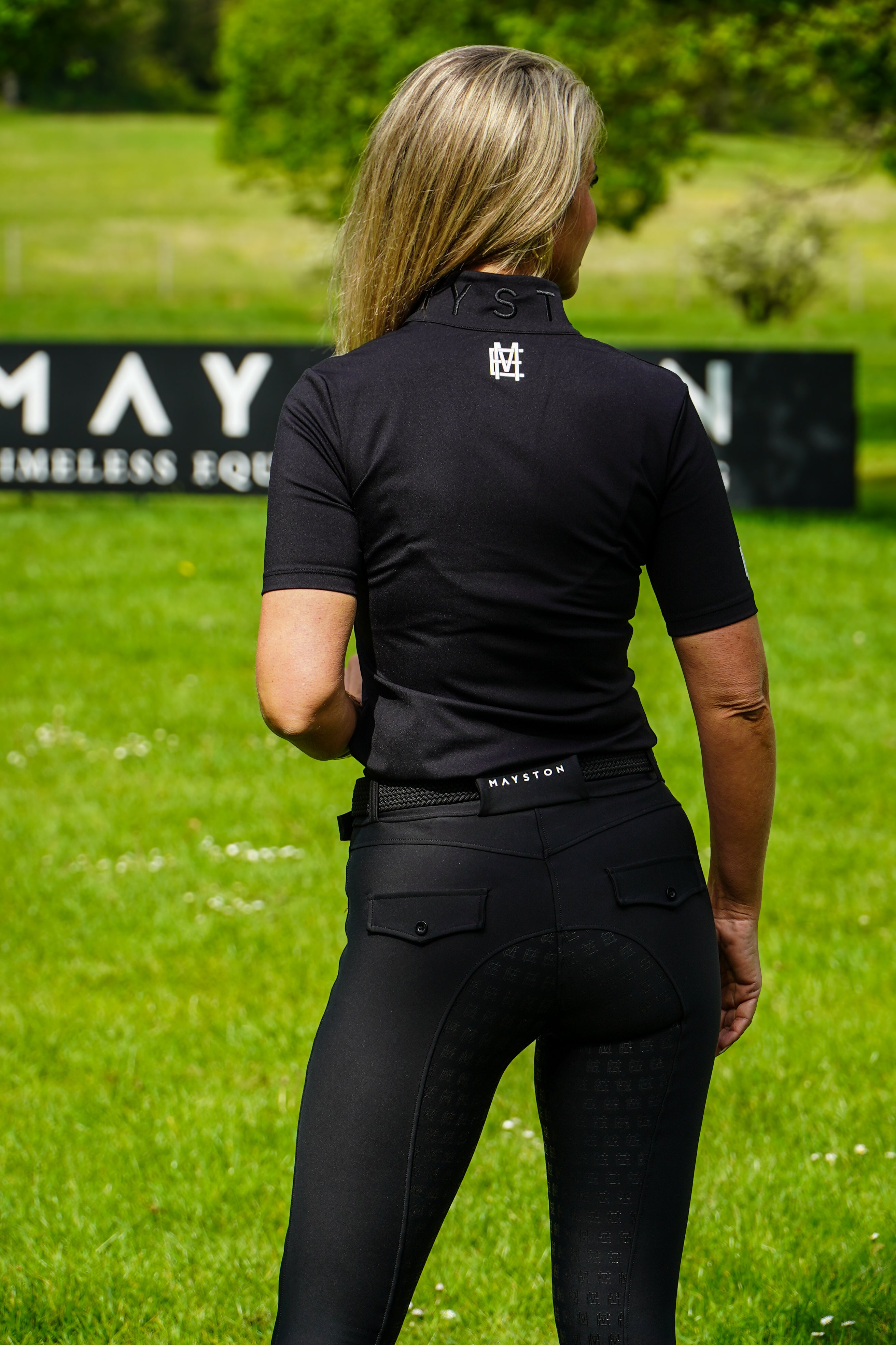 Person wearing a black equestrian shirt and pants on a grassy field with 'May' branding. Broadstone Short sleeve Luxury Sports material base layer short sleeve. Black with subtle white branding colour match embroidery. High collar that's not restrictive. 1/4 zip design
