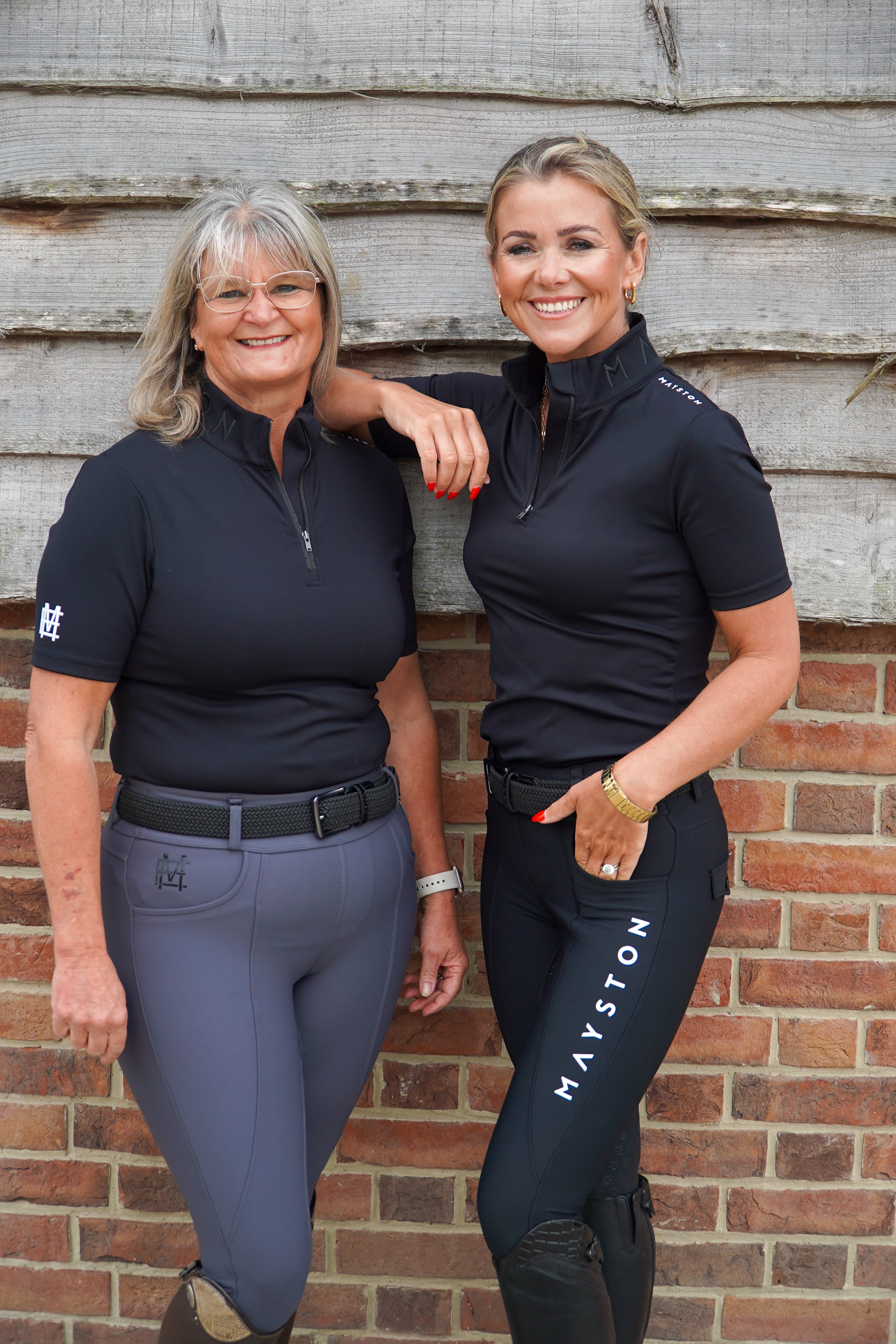 Two women in equestrian attire standing against a brick wall. Broadstone Short sleeve Luxury Sports material base layer short sleeve. Black with subtle white branding colour match embroidery. High collar that's not restrictive. 1/4 zip design