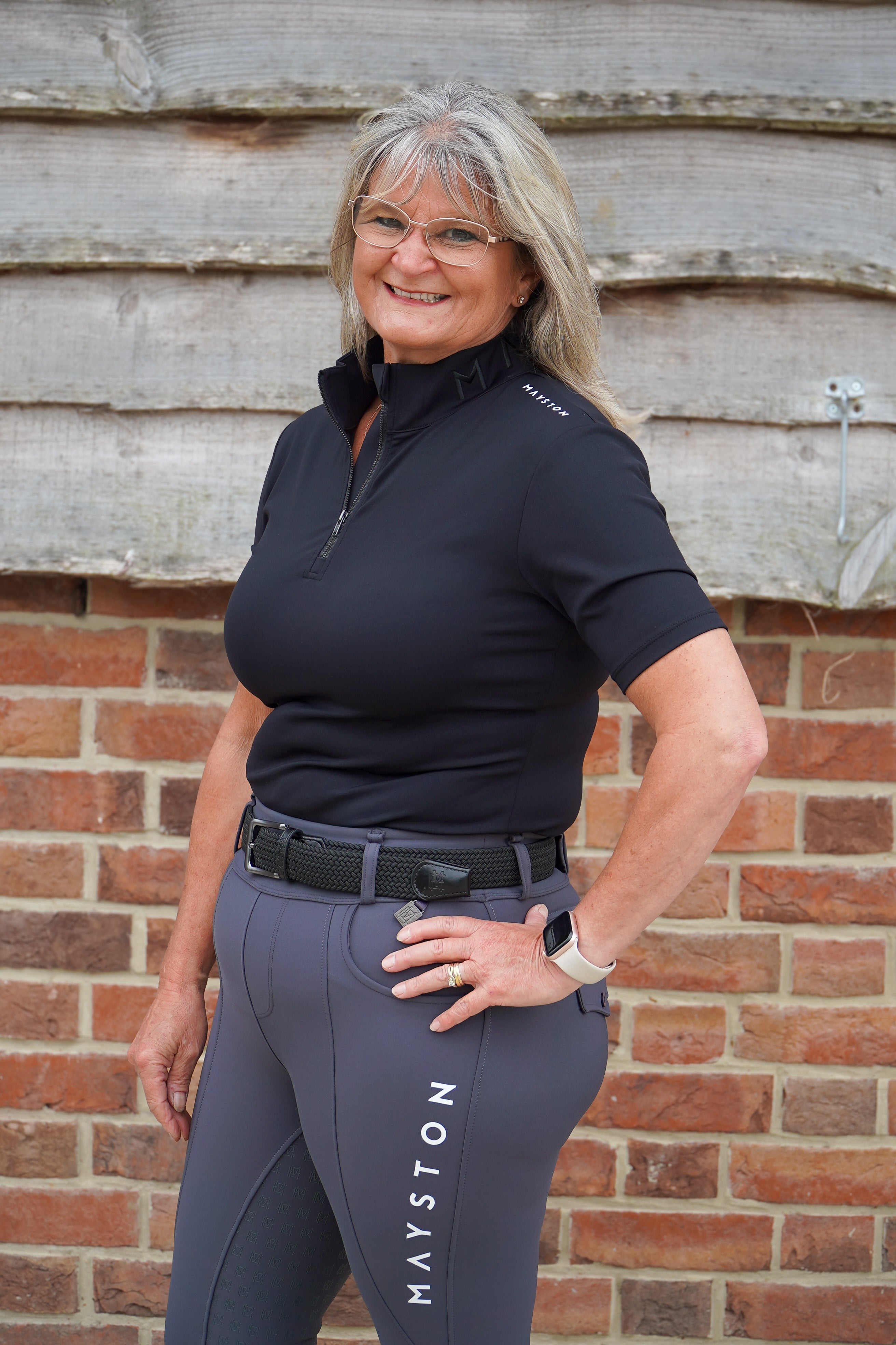 Woman wearing a black polo shirt and grey riding pants with 'Mayston' branding in front of a brick wall. Broadstone Short sleeve Luxury Sports material base layer short sleeve. Black with subtle white branding colour match embroidery. High collar that's not restrictive. 1/4 zip design