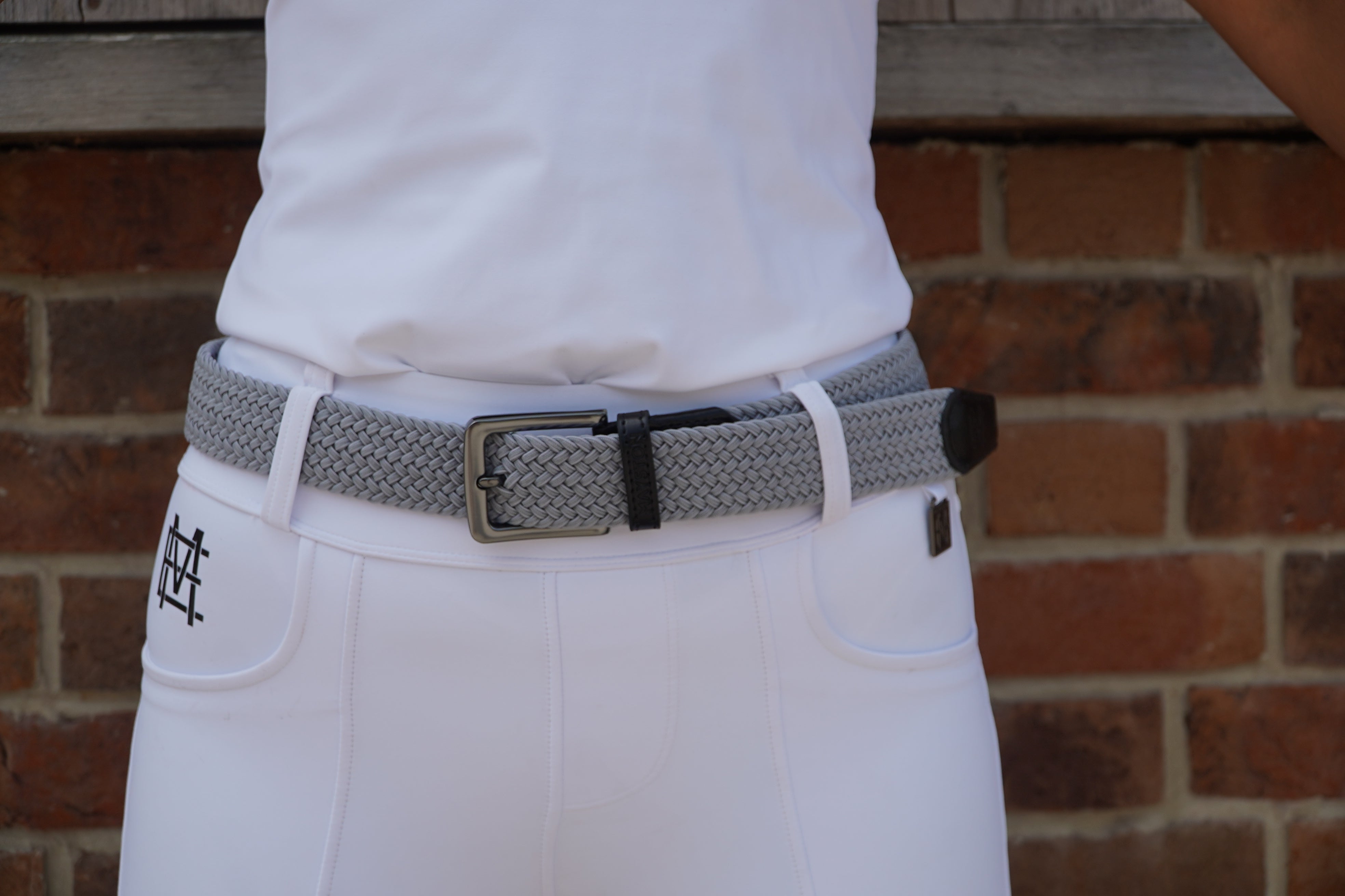 Gray belt with a black buckle on white pants against a brick wall. Elastic stretch quality belt for casual or riding wear