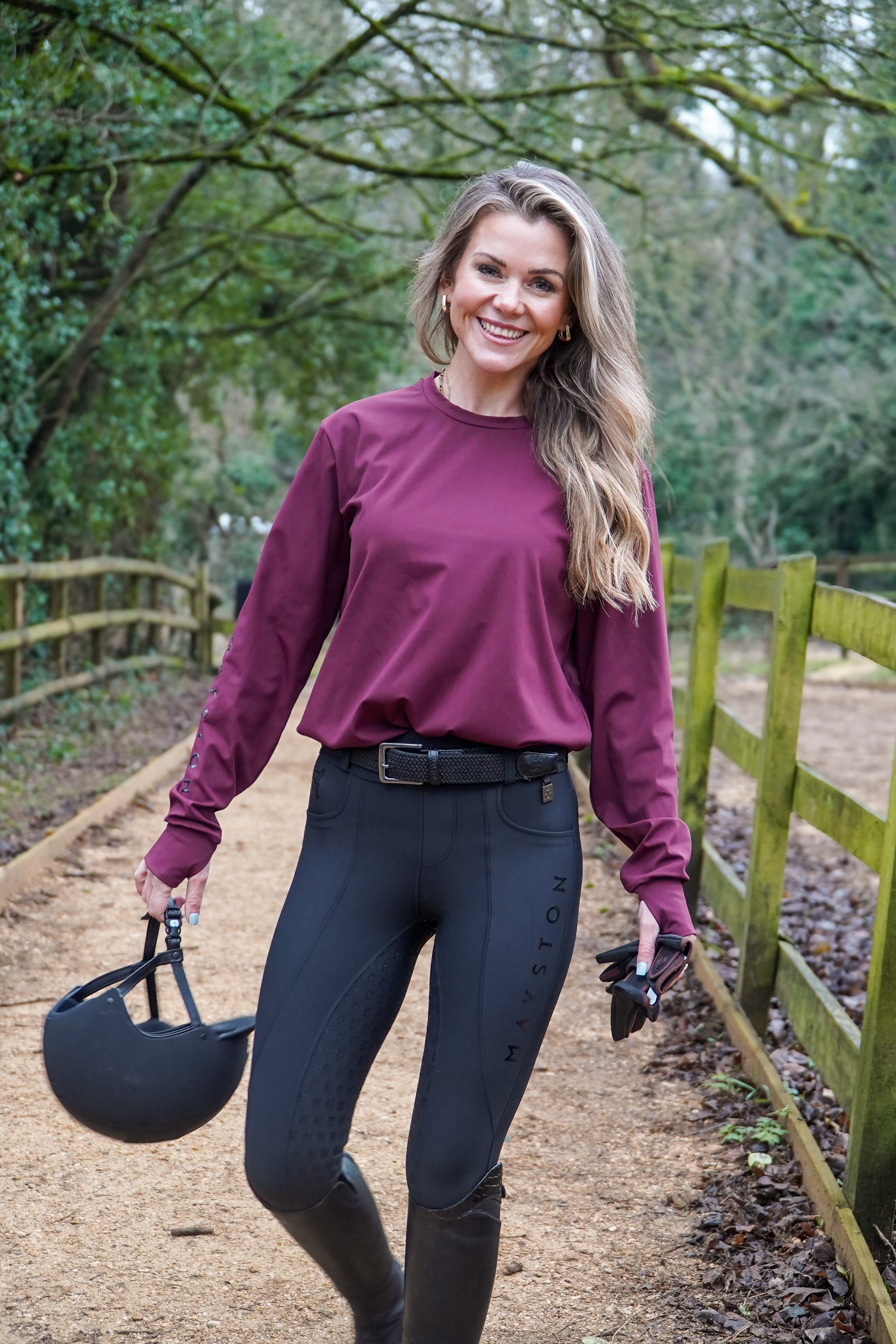 Woman in equestrian attire walking outdoors with a helmet and boots. Deep rich burgundy classic colour. Luxury quality long sleeve t shirt and fabric is specially crafted for active duty, providing flexibility and ease of movement for any busy lifestyle. An ideal choice for a day at the yard, in the saddle, or during your grooming duties, its loose-fitting design guarantees absolute comfort. added thumbholes, Subtle branding sleek design