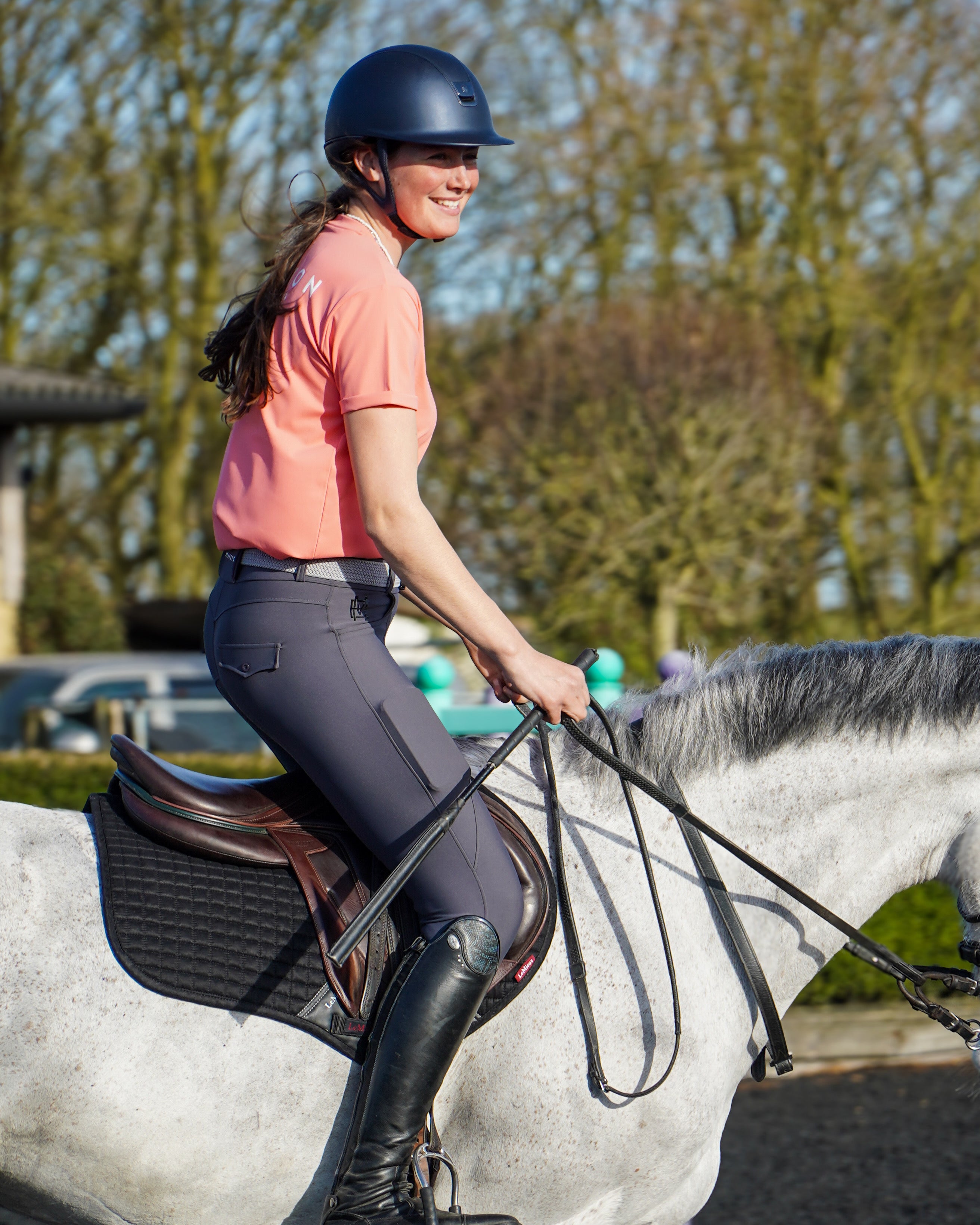 Person riding a horse in an outdoor setting with trees in the background. luxury coral pink quality t shirt and fabric is specially crafted for active duty, providing flexibility and ease of movement for any busy lifestyle. An ideal choice for a day at the yard, in the saddle, or during your grooming duties, its loose-fitting design guarantees complete freedom.