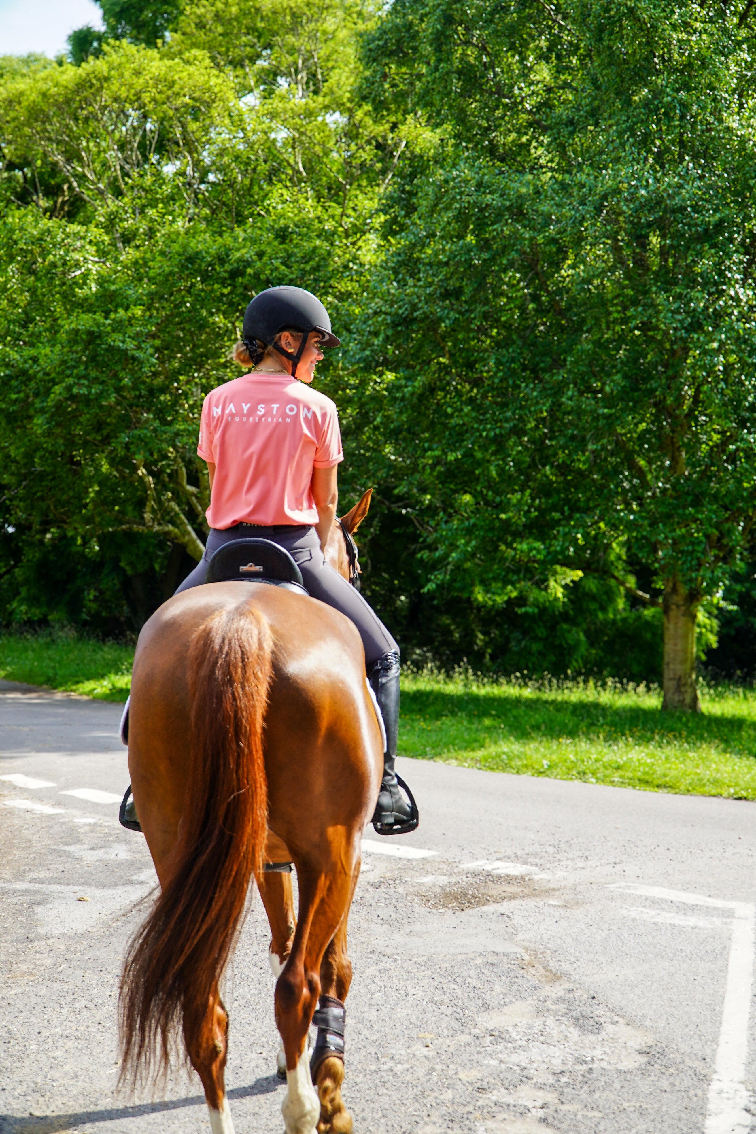 Person riding a horse on a paved path with trees in the background. luxury quality pink t shirt and fabric is specially crafted for active duty, providing flexibility and ease of movement for any busy lifestyle. An ideal choice for a day at the yard, in the saddle, or during your grooming duties, its loose-fitting design guarantees complete freedom.