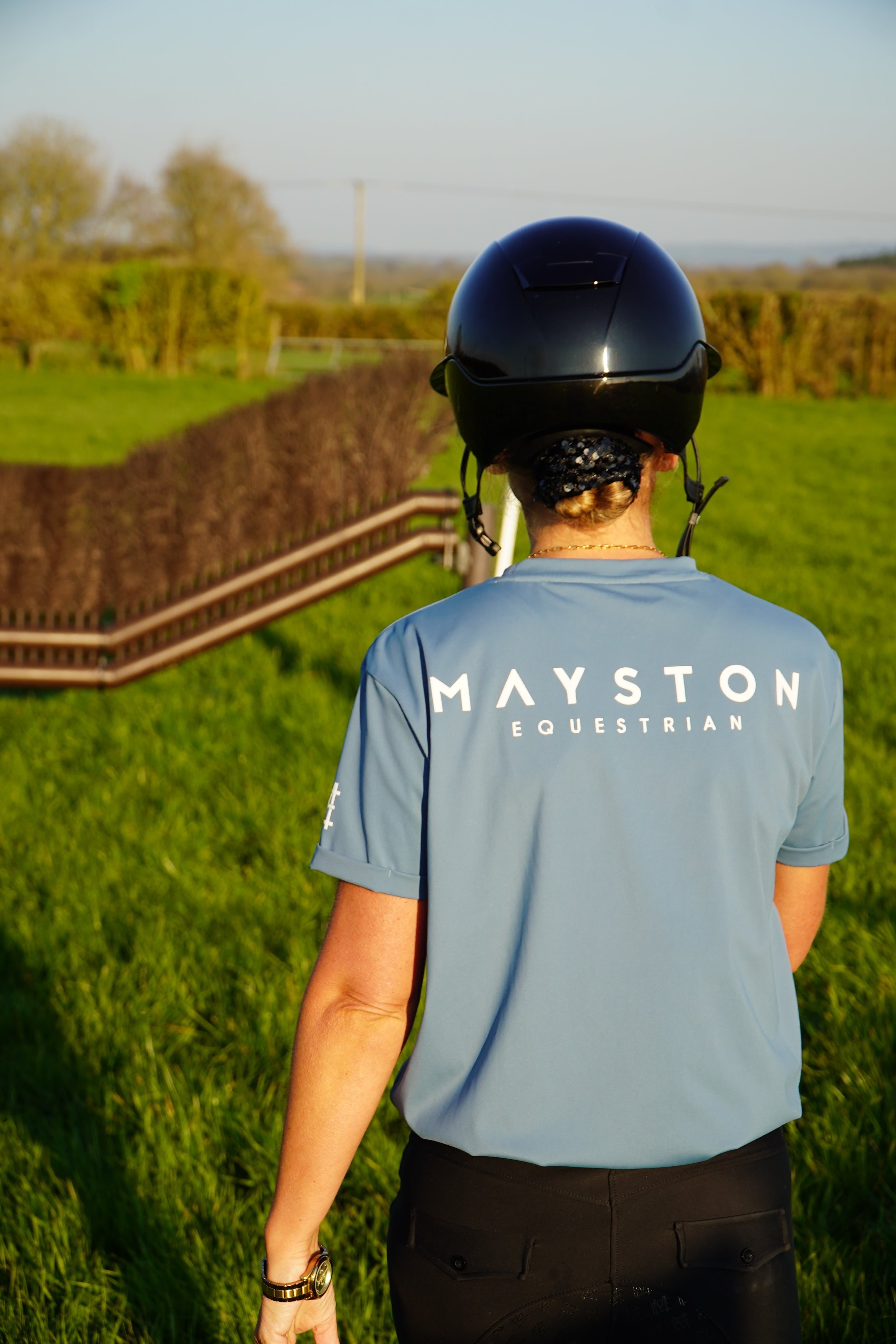 Person wearing a blue shirt with 'MAYSTON' branding, standing on grass with a path and trees in the background. A person wearing a blue t-shirt with the text 'MAYSTON' on the back, standing in an outdoor setting. luxury quality t shirt and fabric is specially crafted for active duty, providing flexibility and ease of movement for any busy lifestyle. An ideal choice for a day at the yard, in the saddle, or during your grooming duties, its loose-fitting design guarantees complete freedom. high gloss white bra