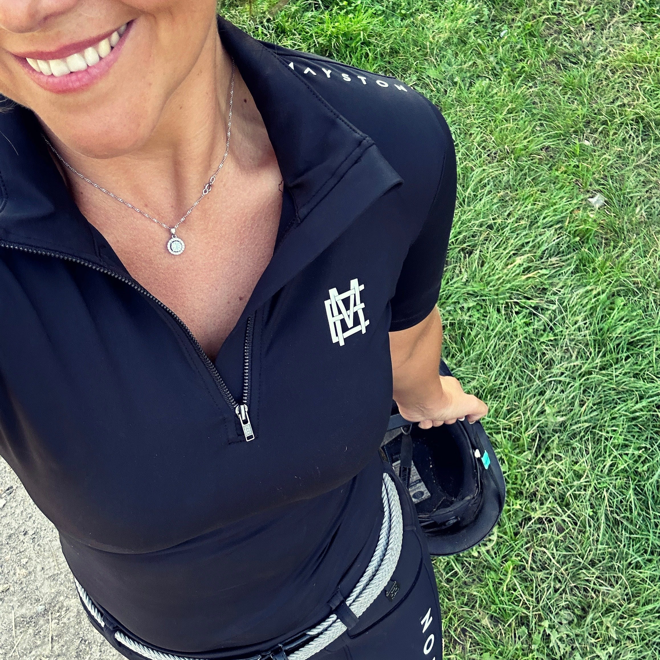 Sleek silhouette baselayer top subtle Mayston branding, maximum coverage without compromising style, next-level performance with a bold, minimalist look. Moisture-wicking, breathable fabric with four-way stretch, it keeps you cool and comfortable. Training, Everyday or competition wear. Short Sleeve 1/4 zip with collar. classic black with grey and white branding. Woman Standing in equestrian wear