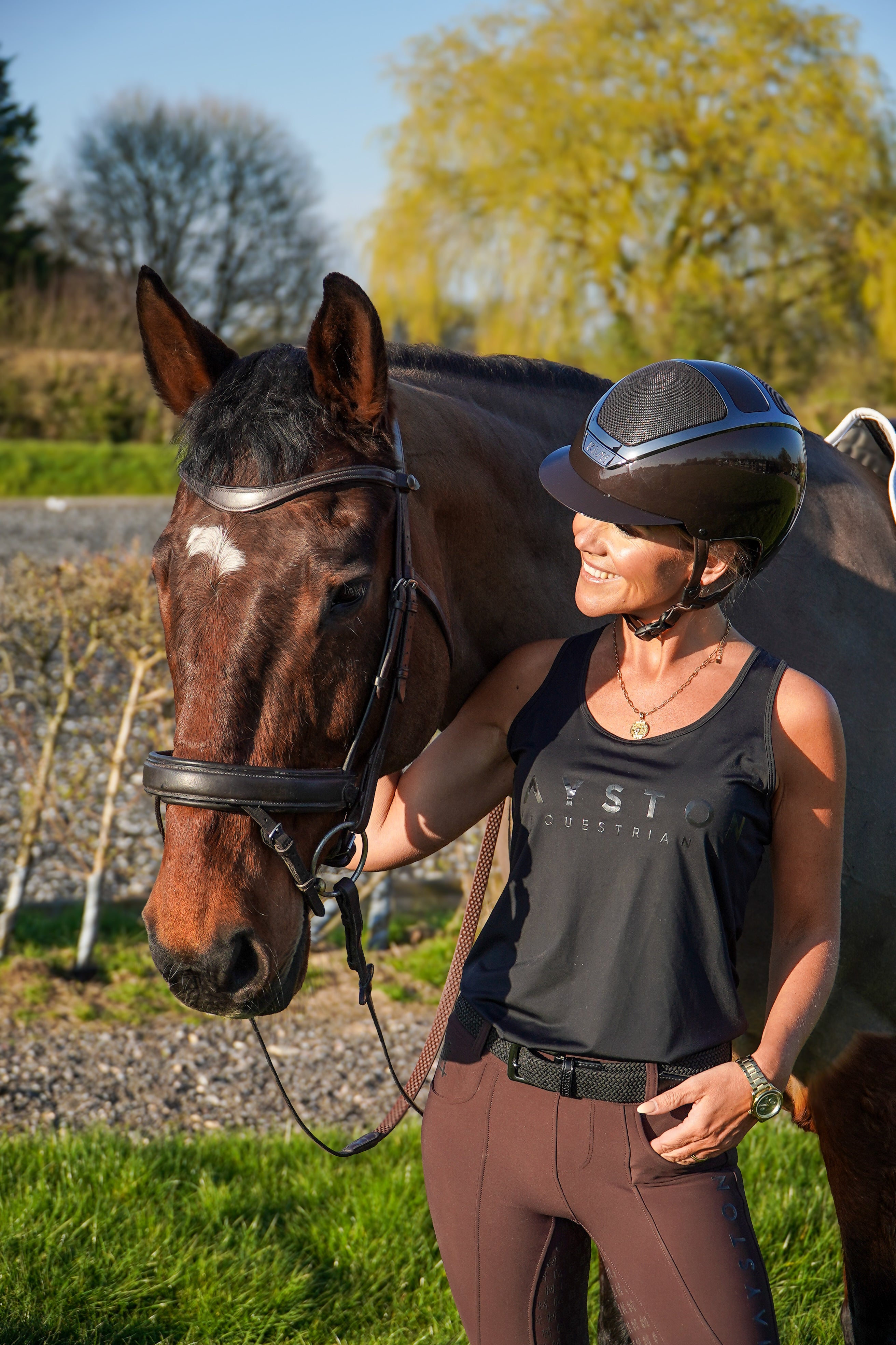 Woman in equestrian gear standing next to a horse in an outdoor setting. Radipole Racerback Vest, crafted from lightweight, high-quality sports material for effortless comfort and performance. Designed to move with you, it offers breathability and freedom of motion, making it perfect for riding, training, or everyday wear. Finished with bold white high-gloss Mayston silicone branding on the chest and a sleek logo at the rear, this black vest combines sporty style with signature Mayston.