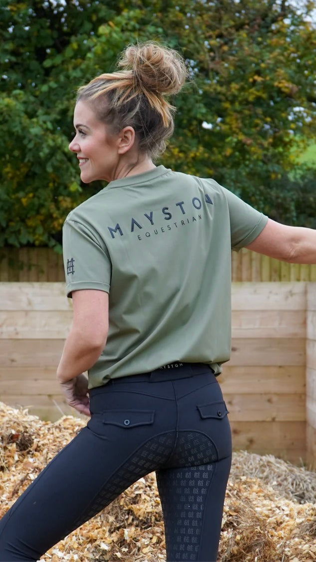 A person wearing a green t-shirt with the brand name 'DURLSTON' across the back, riding apparel, and black leggings with a silicone print detail on the back. luxury quality t shirt and fabric is specially crafted for active duty, providing flexibility and ease of movement for any busy lifestyle. An ideal choice for a day at the yard, in the saddle, or during your grooming duties, its loose-fitting design guarantees complete freedom.