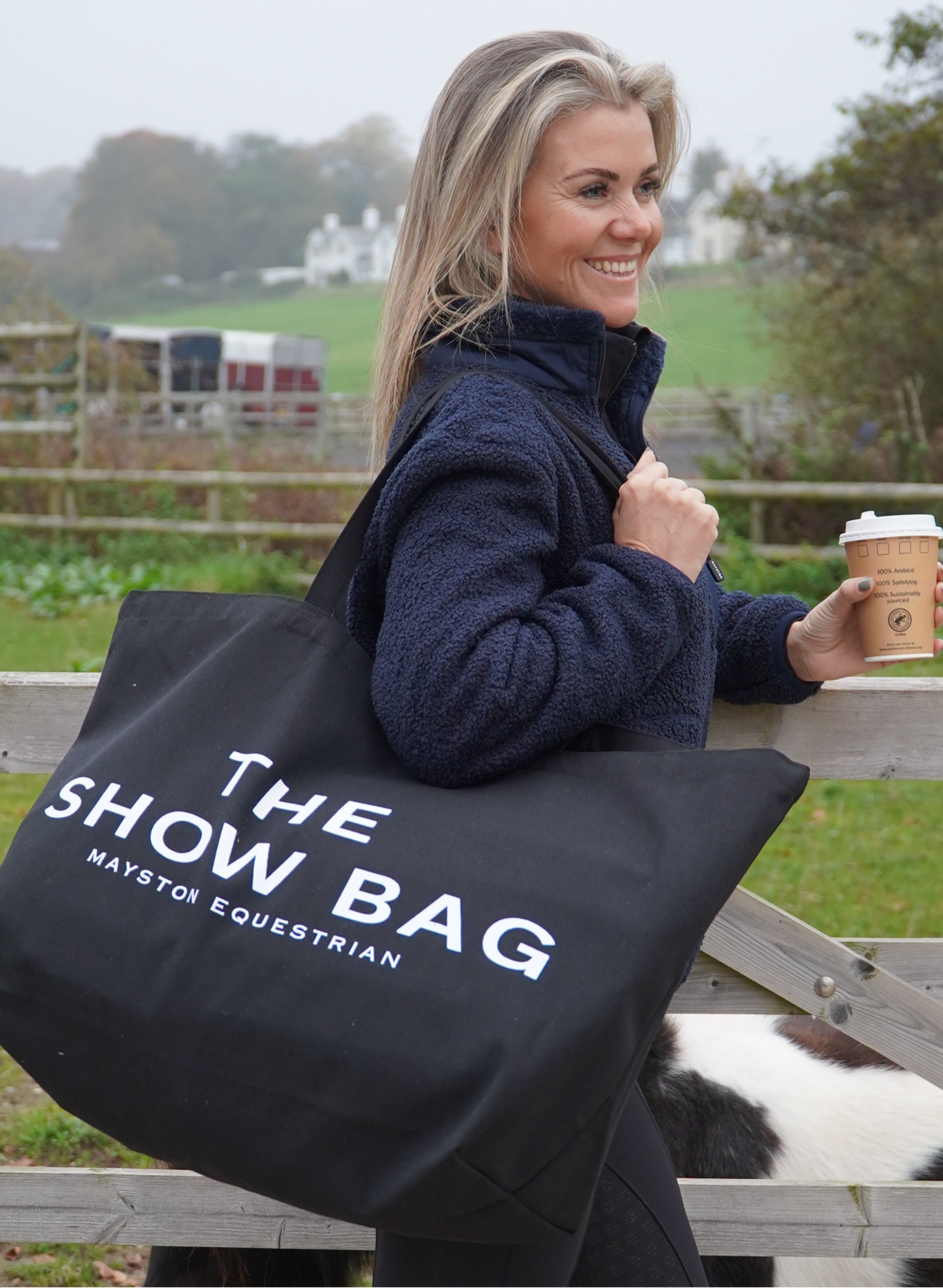 A woman carrying a black tote bag with the text 'THE SHOW BAG' printed on it, in a equestrian setting.