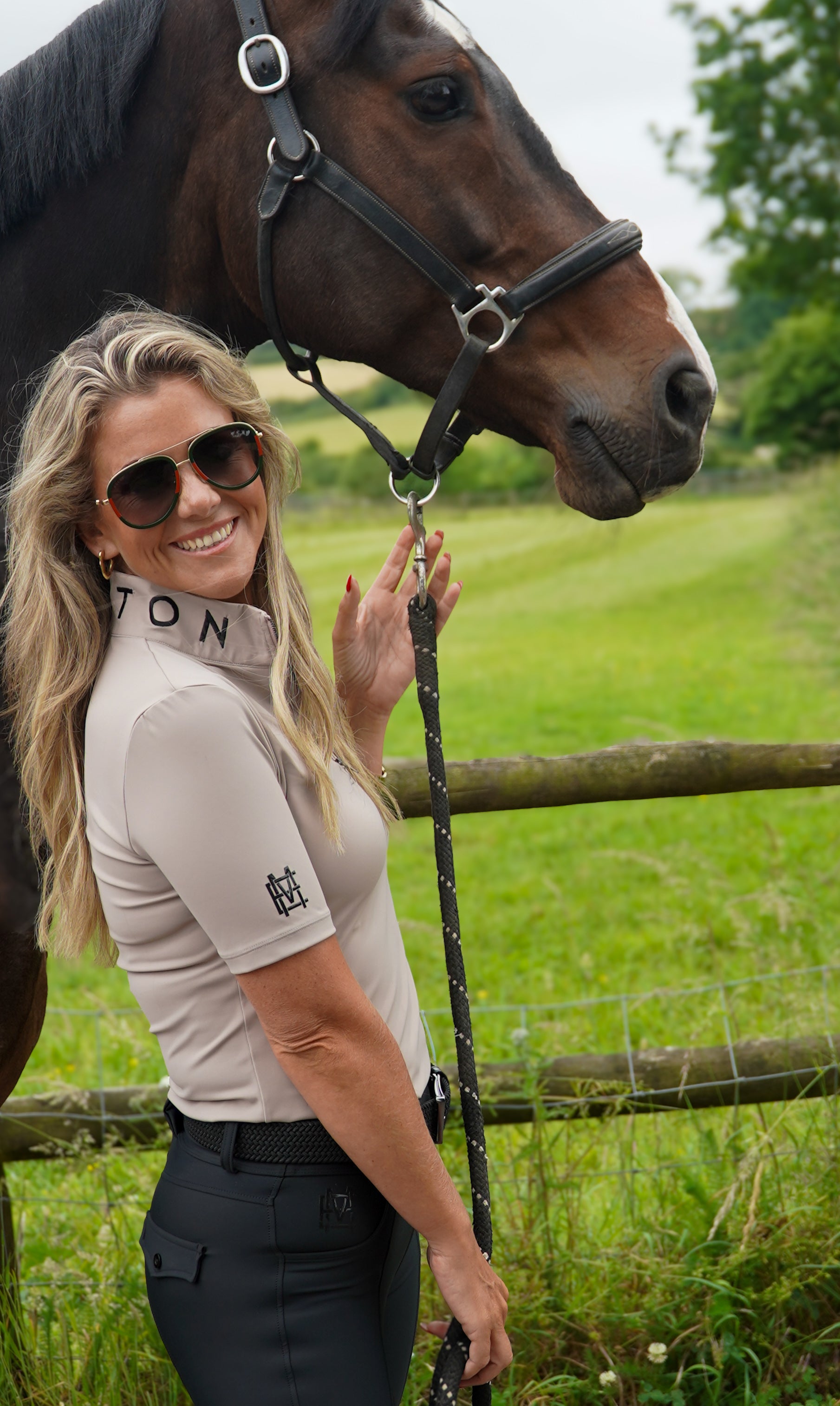 Woman in equestrian attire standing next to a horse in a grassy field. Crafted from our soft, breathable performance fabric, it keeps you cool without compromising on style. Finished in taupe, with MAYSTON embroidery at the collar and black silicone branding on the sleeve, it’s a versatile piece that transitions effortlessly from yard to everyday wear.