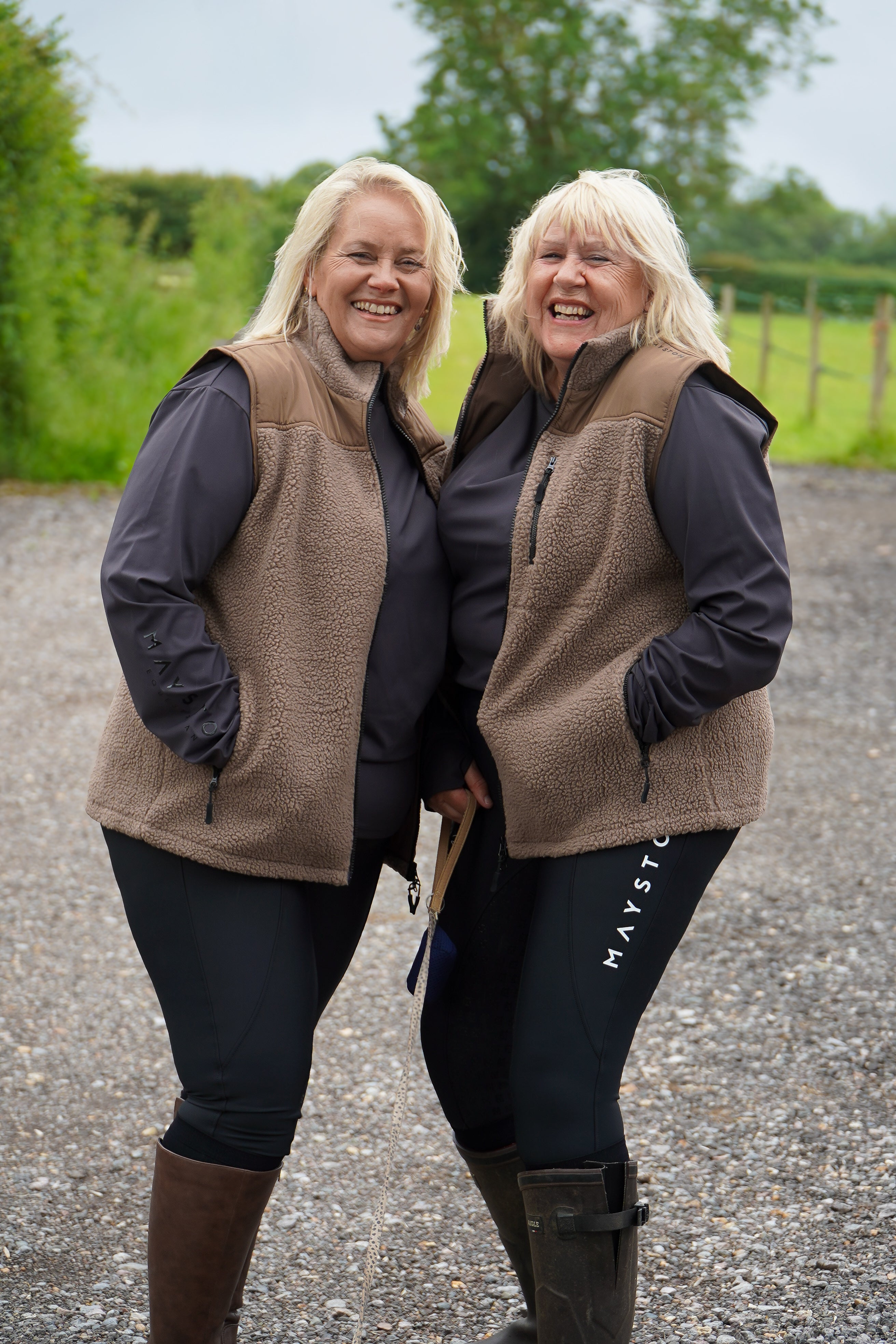 Two women wearing matching brown vests and dark pants standing outdoors on a gravel path with greenery in the background. Luxury teddy Fleece Gilet Waistcoat. Quality embroidery. multiple pockets. Subtle branding, elegant classic design. Stretch lining