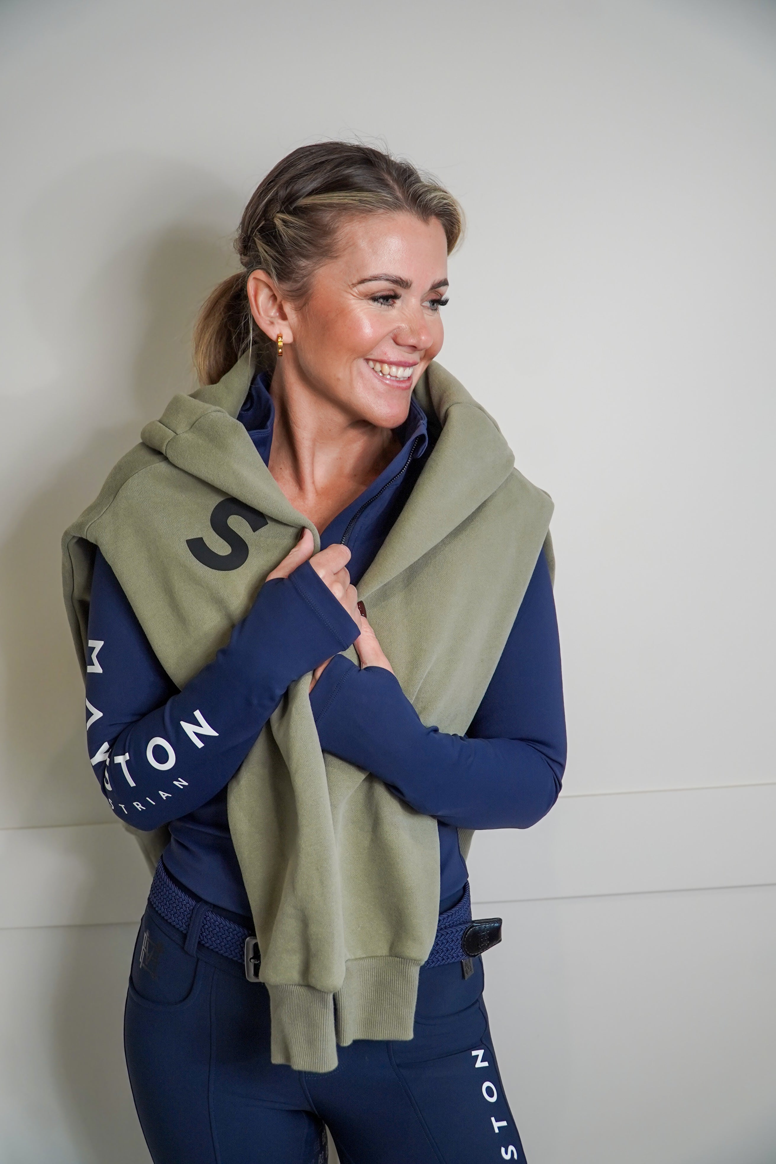 Woman wearing a navy jacket with branding and an olive green hoodie jumper over it, standing against a plain background. High quality luxurious 100% cotton. wrap around bold logo. soft, cozy and warm. Hoodie, jumper sweatshirt. everyday wear. casual and smart