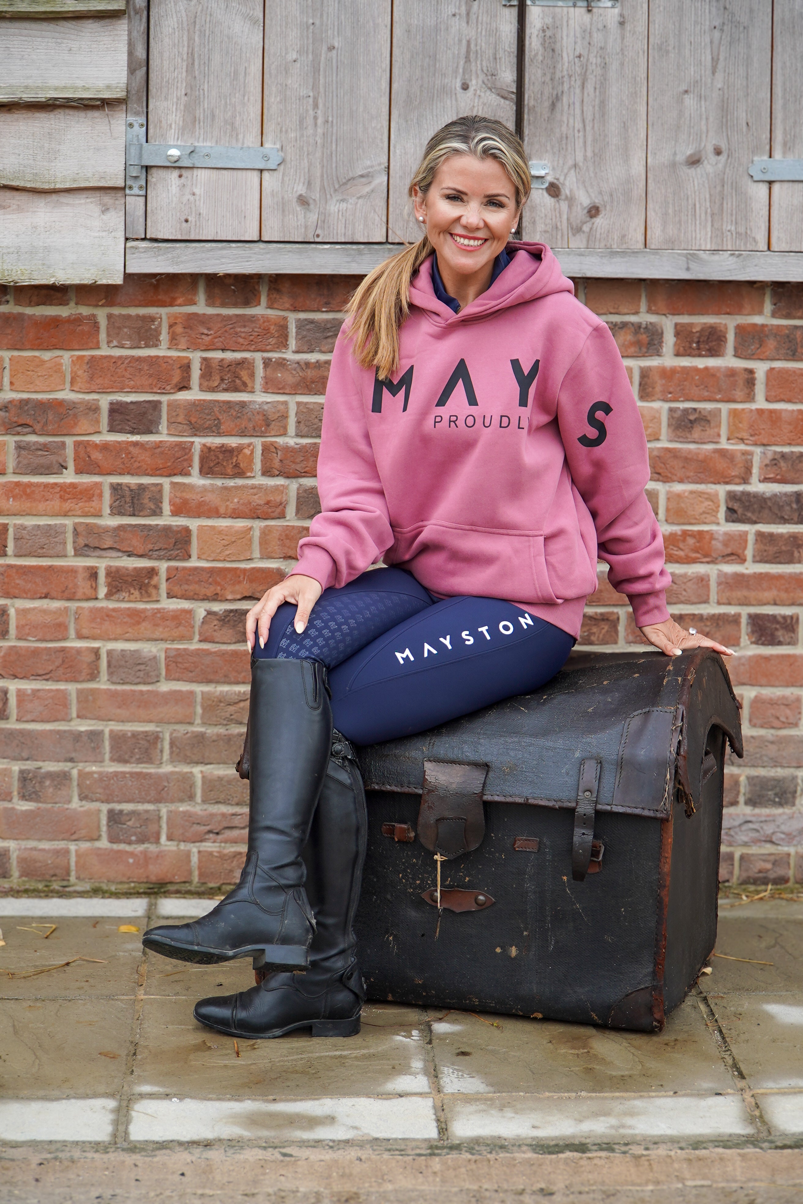 Woman in pink hoodie and blue breeches sitting on a vintage suitcase with a brick wall background. High quality luxurious 100% cotton. wrap around bold logo. soft, cozy and warm. Hoodie, jumper sweatshirt. everyday wear. casual and smart