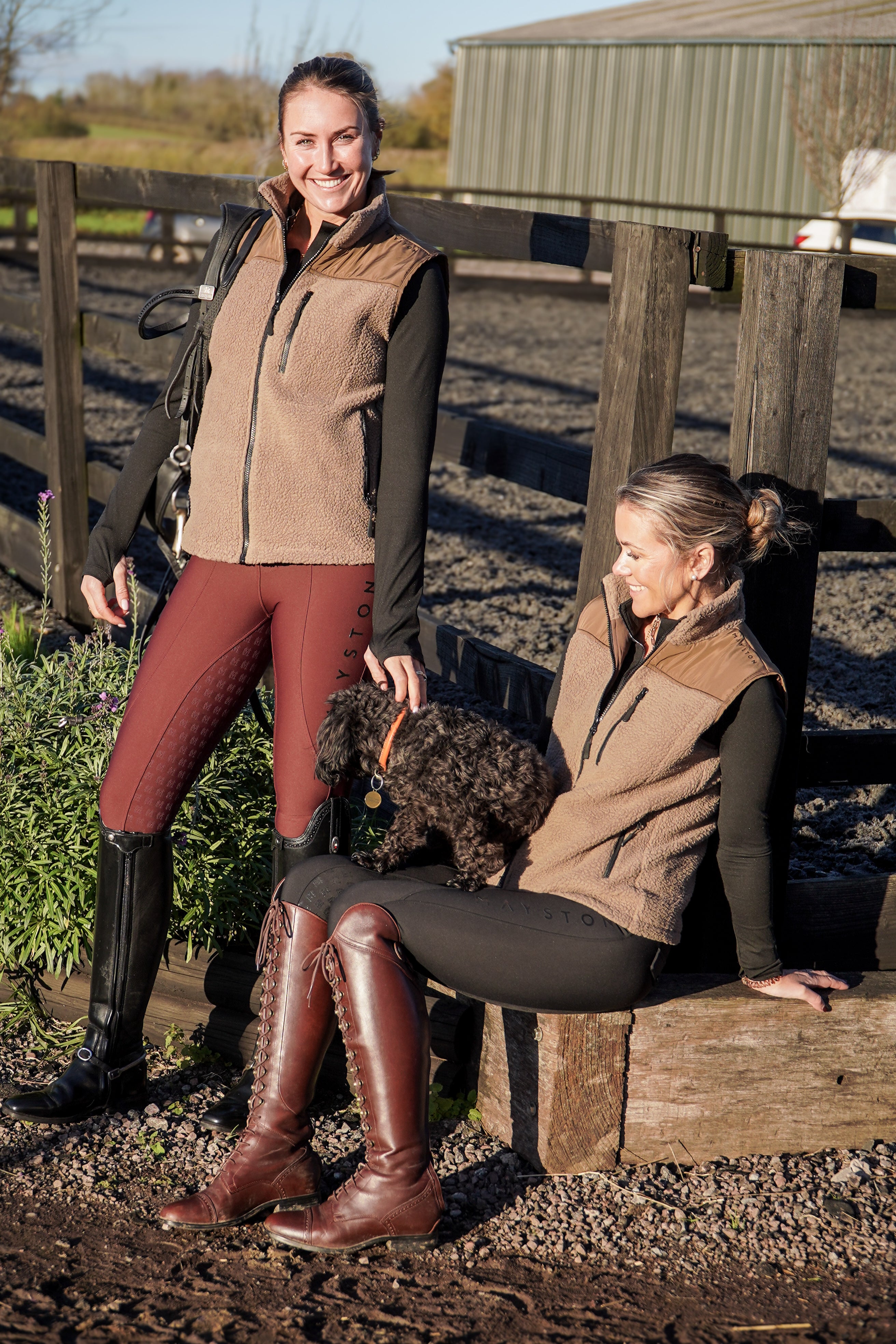 Two women in equestrian attire with a dog on a wooden bench outdoors. luxury fleece gilet with quality embroidery