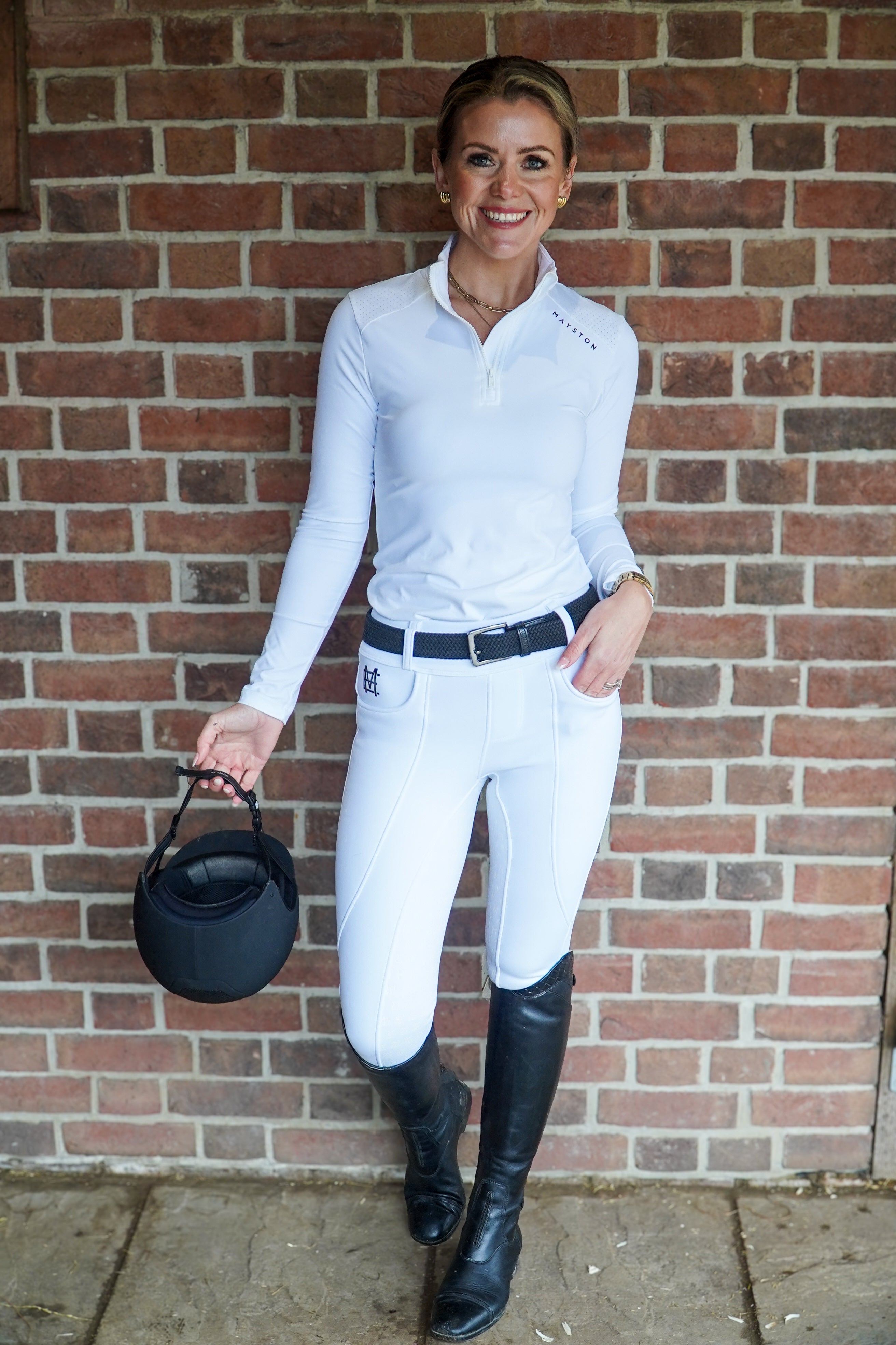 Woman in equestrian attire standing against a brick wall. stretch white breeches, winter fleece lined. full seat silicone