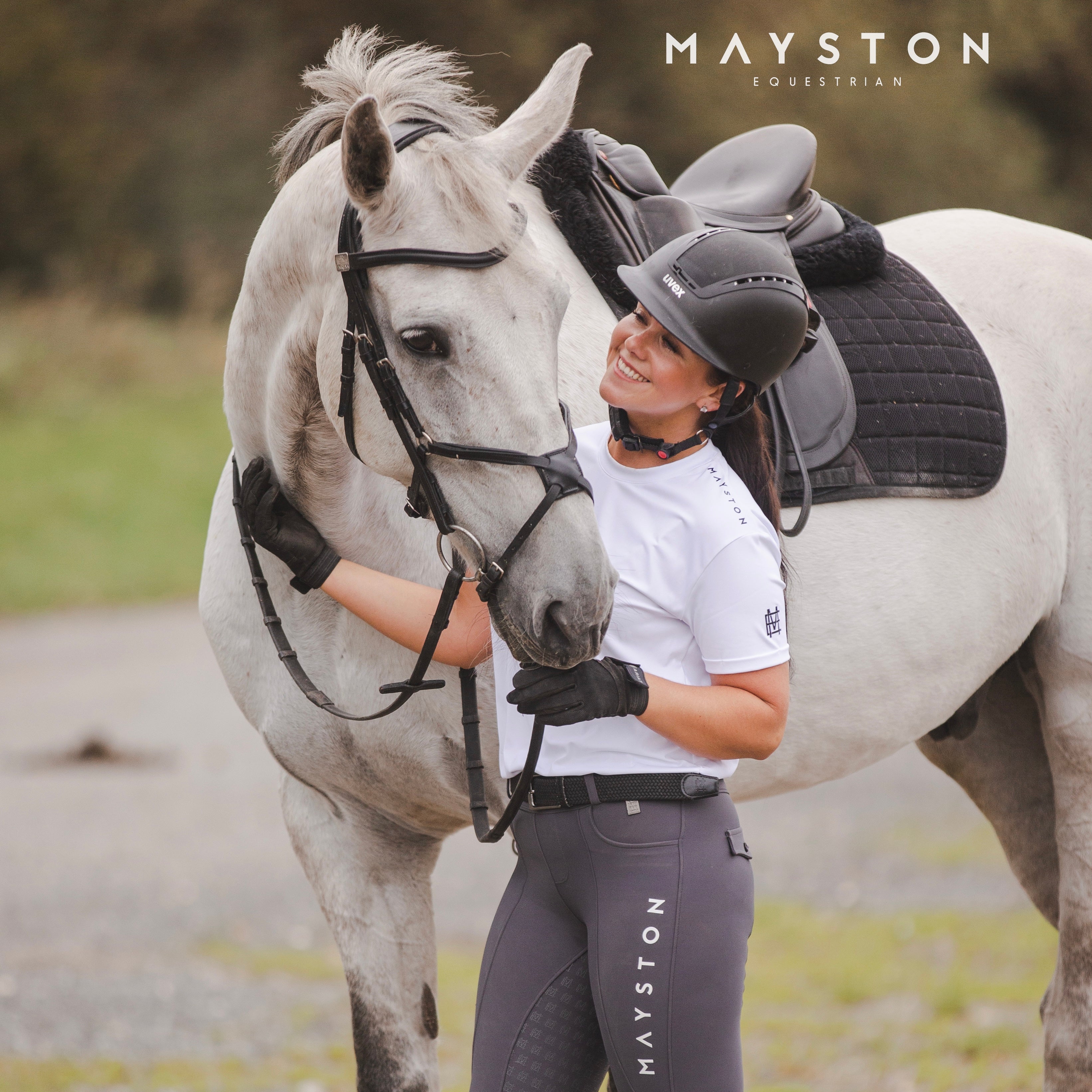 A woman facing away from the camera wears a white t-shirt with the brand name 'MAYSTON EQUESTRIAN' printed across the back, in front, and on the sleeves. She has her hair styled in a bun and is wearing black pants. luxury quality t shirt and fabric is specially crafted for active duty, providing flexibility and ease of movement for any busy lifestyle. An ideal choice for a day at the yard, in the saddle, or during your grooming duties, its loose-fitting design guarantees complete freedom.