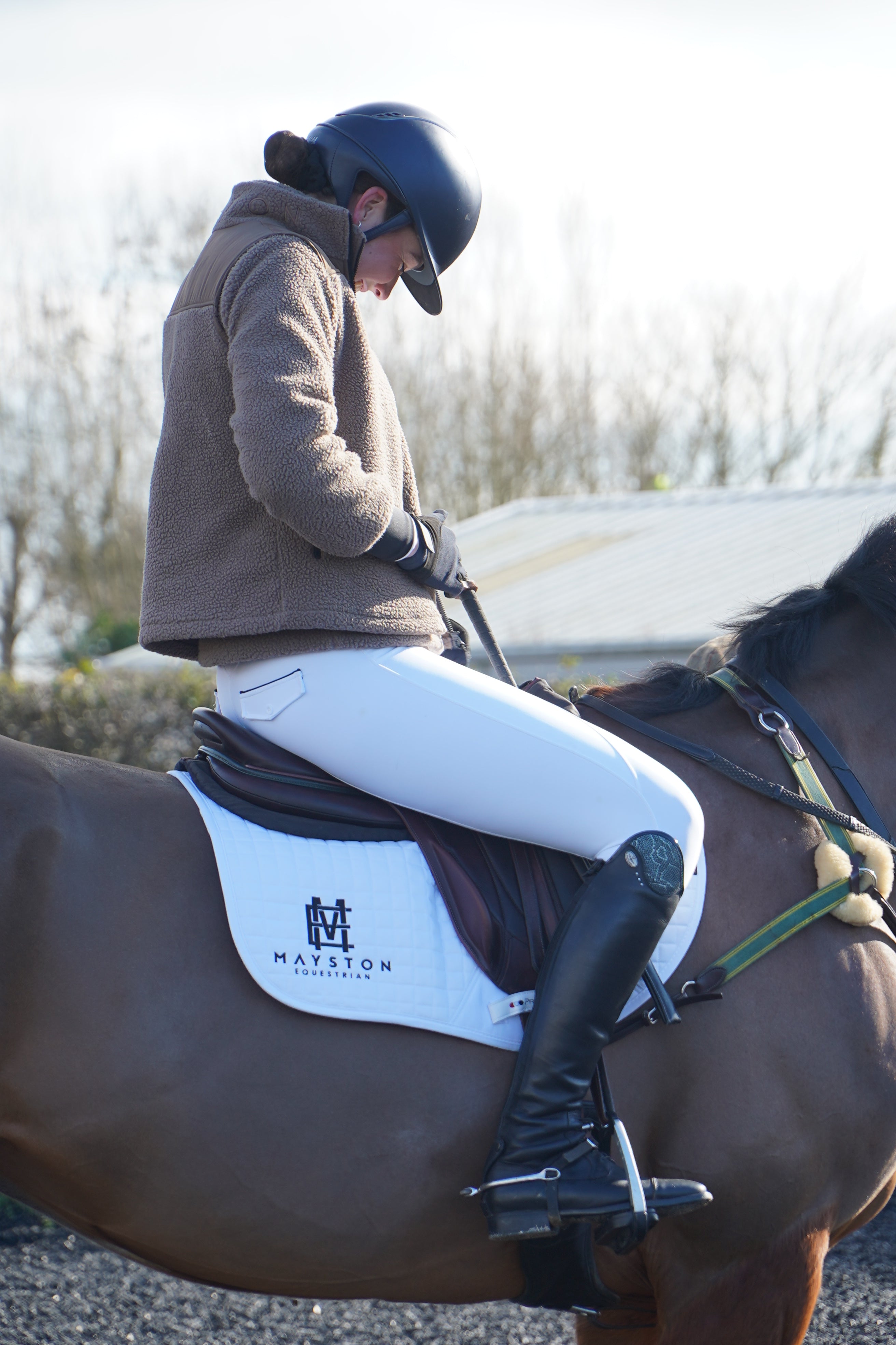 Person riding a horse with a saddle and bridle, standing by a body of water. Premium white competition breeches with a Full Silicone seat. Stretch high waisted Jodhpurs. Deep pockets, supportive, comfortable and compression quality fabric. Elegant detailing with subtle branding.