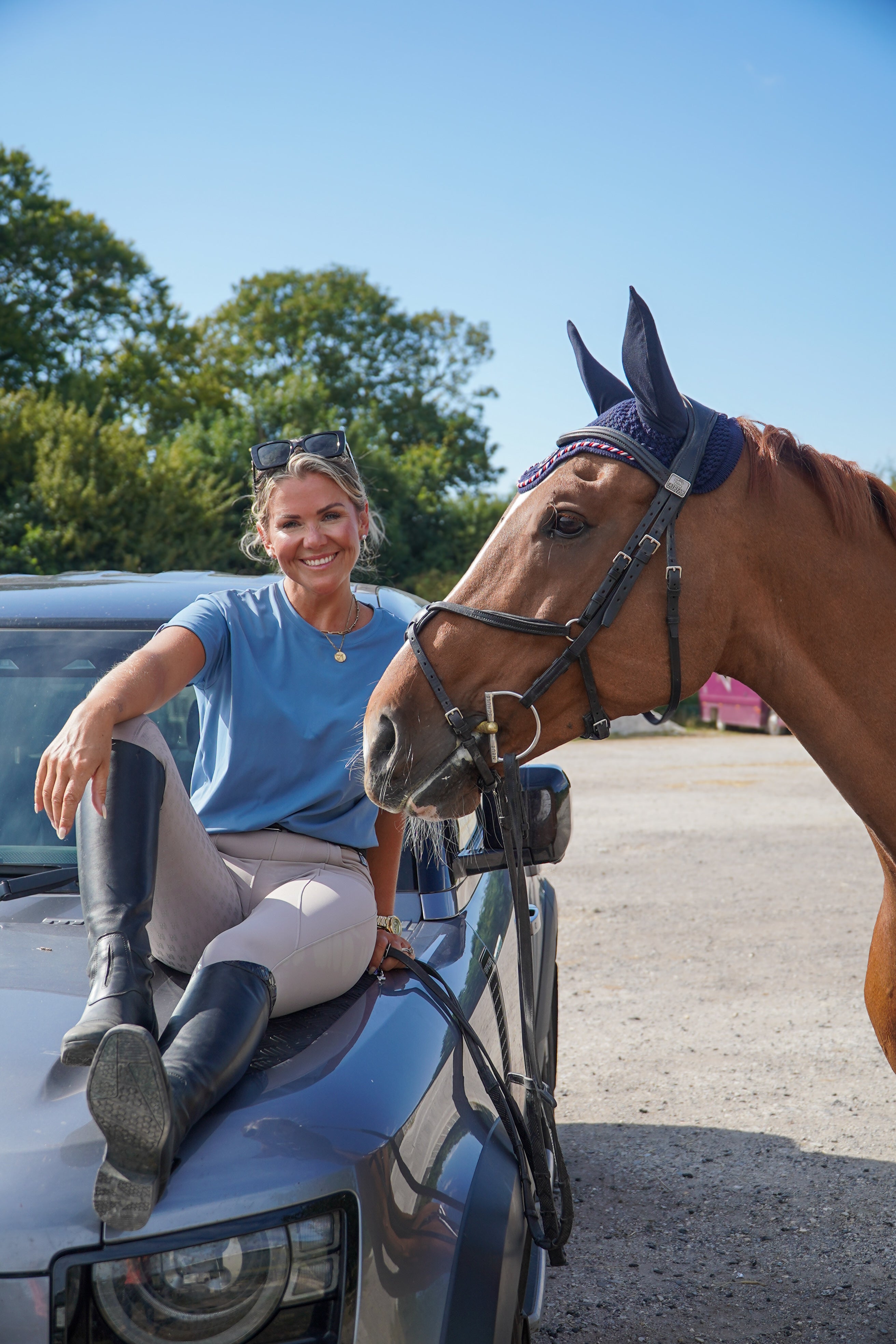 Woman sitting on a car with a horse next to her, outdoors. luxury quality t shirt and fabric is specially crafted for active duty, providing flexibility and ease of movement for any busy lifestyle. An ideal choice for a day at the yard, in the saddle, or during your grooming duties, its loose-fitting design guarantees complete freedom.