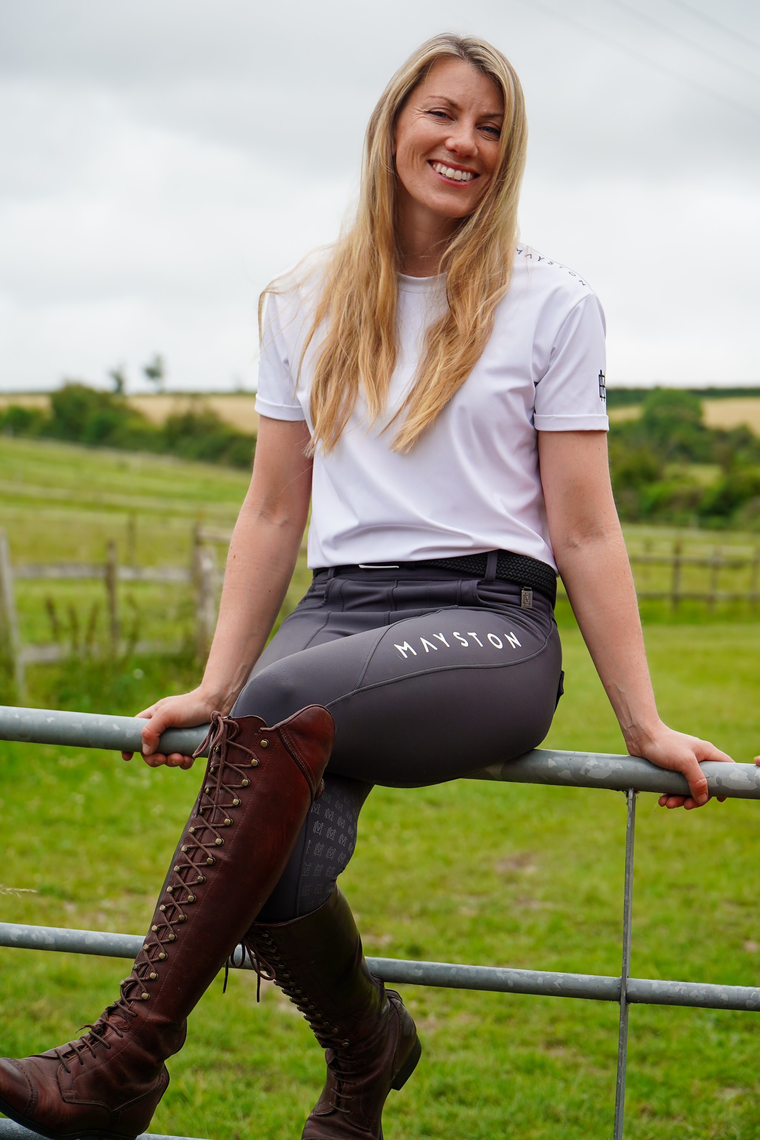 Woman in equestrian attire sitting on a fence in a field. Premium Grey breeches with a Full Silicone seat. Stretch high waisted Jodhpurs. Deep pockets, supportive, comfortable and compression quality fabric. Elegant detailing with subtle branding.