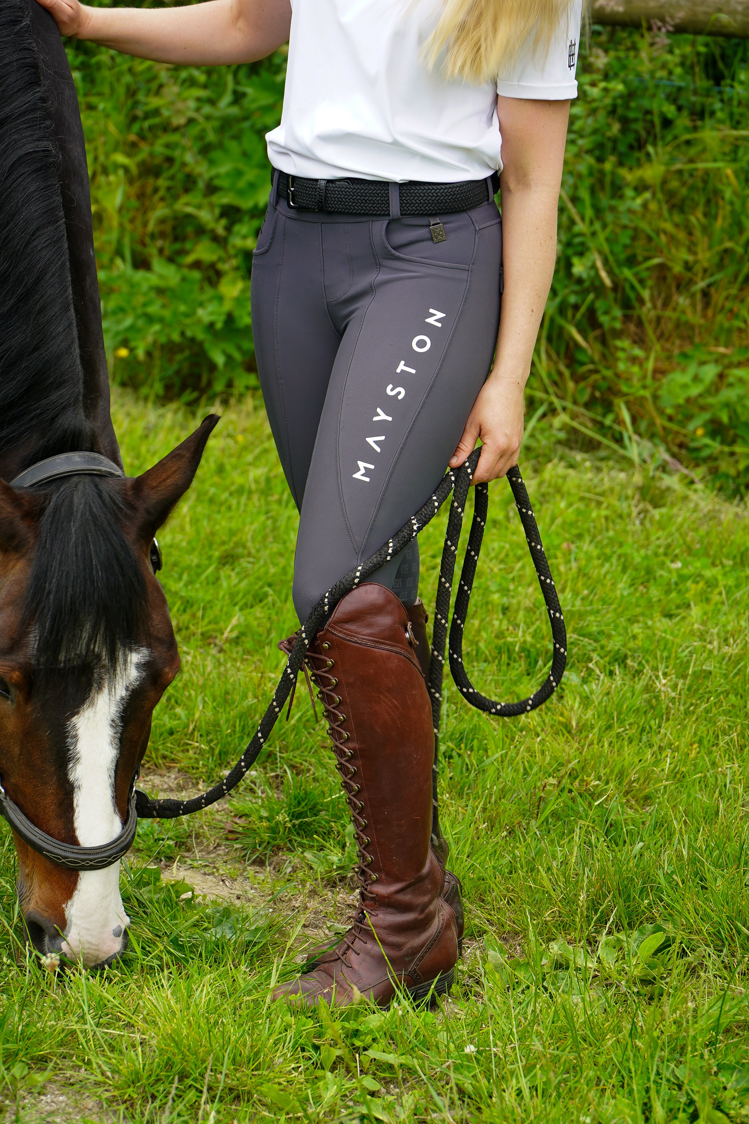 Person in equestrian attire standing next to a horse in a grassy field. Premium Grey breeches with a Full Silicone seat. Stretch high waisted Jodhpurs. Deep pockets, supportive, comfortable and compression quality fabric. Elegant detailing with subtle branding.