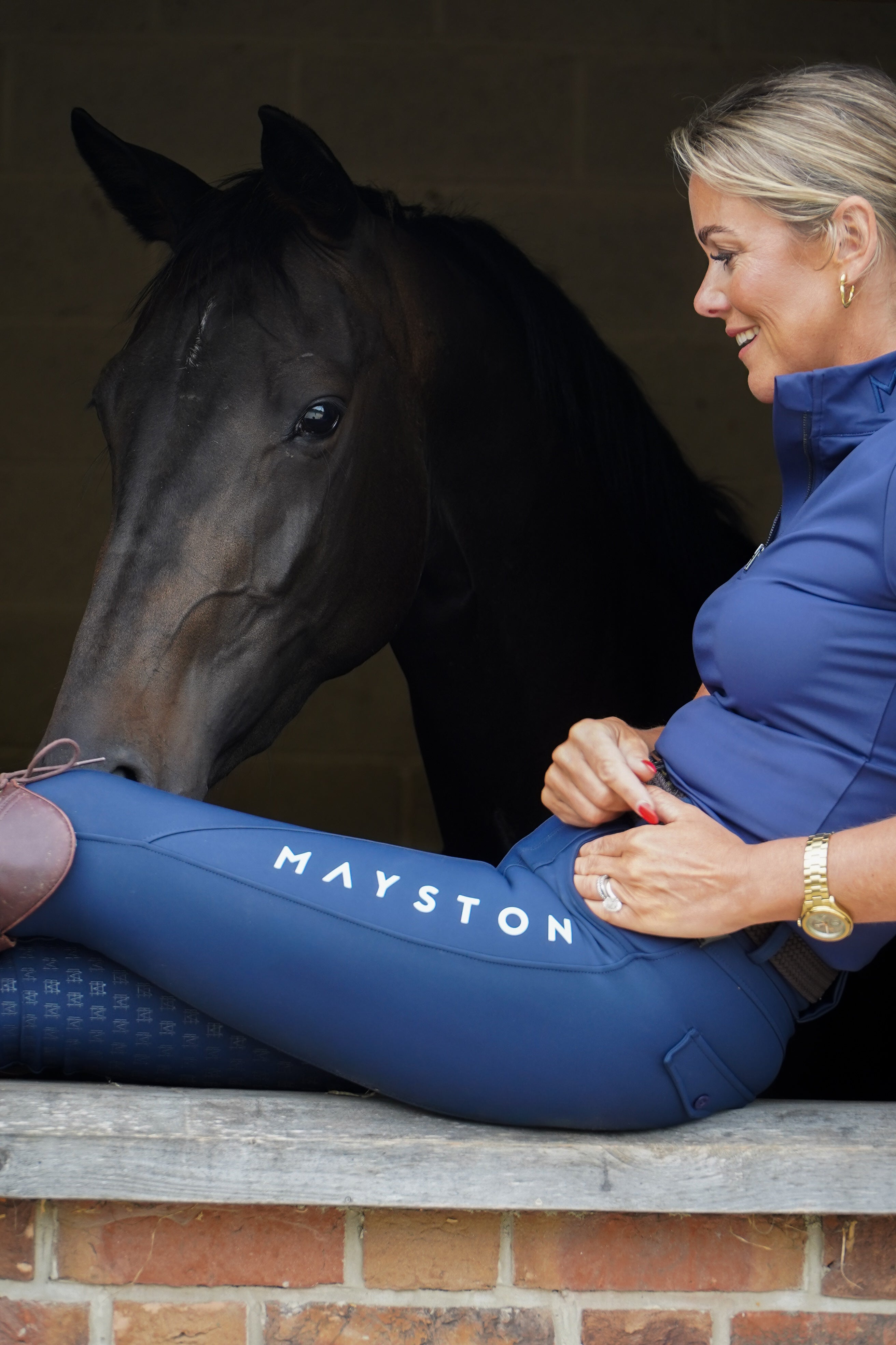 Woman in blue equestrian attire sitting next to a horse with 'Mayston' Premium navy breeches with a Full Silicone seat. Stretch high waisted Jodhpurs. Deep pockets, supportive, comfortable and compression quality fabric. Elegant detailing with subtle branding. high gloss white brandingbranding.