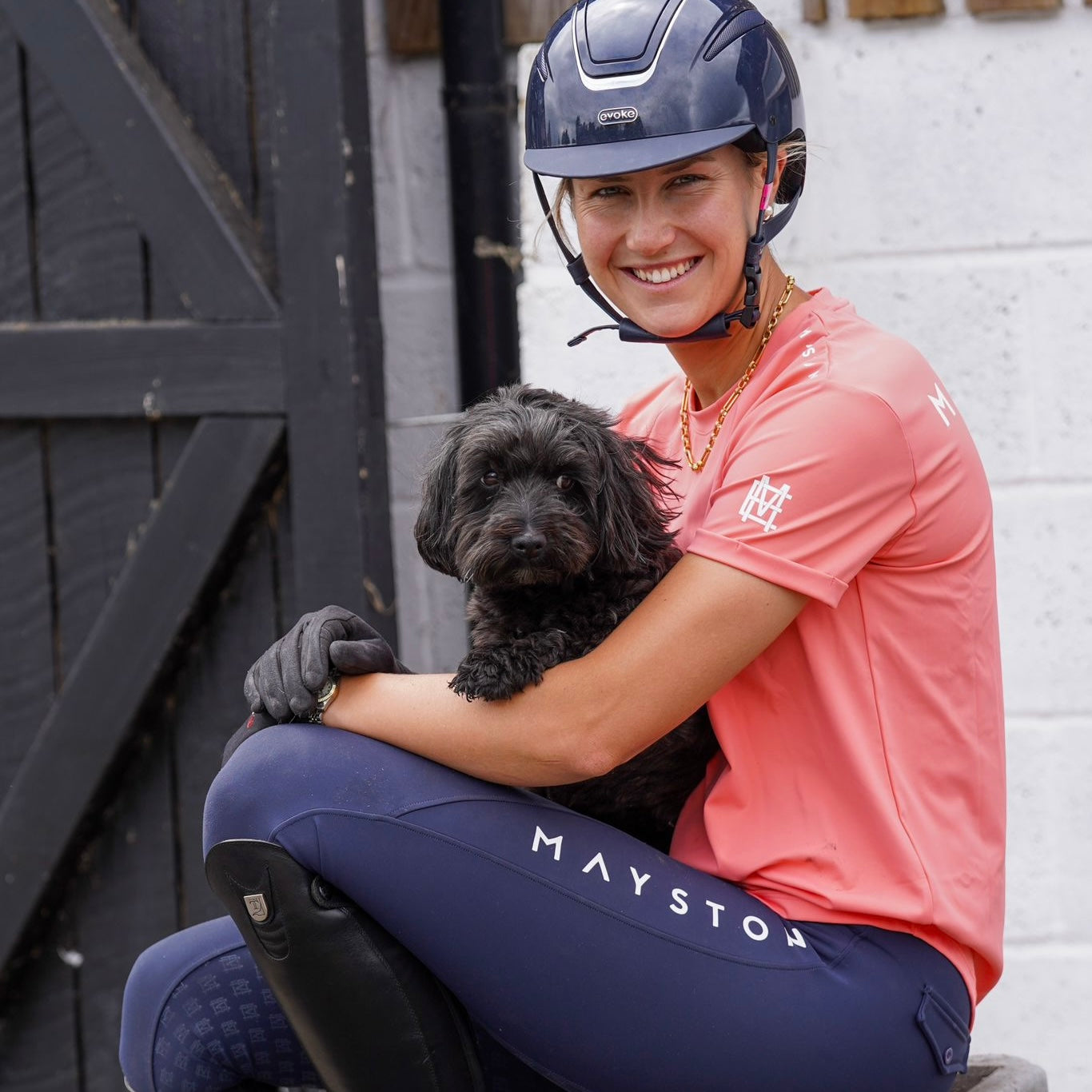 A person wearing a coral pink long-sleeved equestrian t-shirt holding a black dog luxury quality t shirt and fabric is specially crafted for active duty, providing flexibility and ease of movement for any busy lifestyle. An ideal choice for a day at the yard, in the saddle, or during your grooming duties, its loose-fitting design guarantees complete freedom.