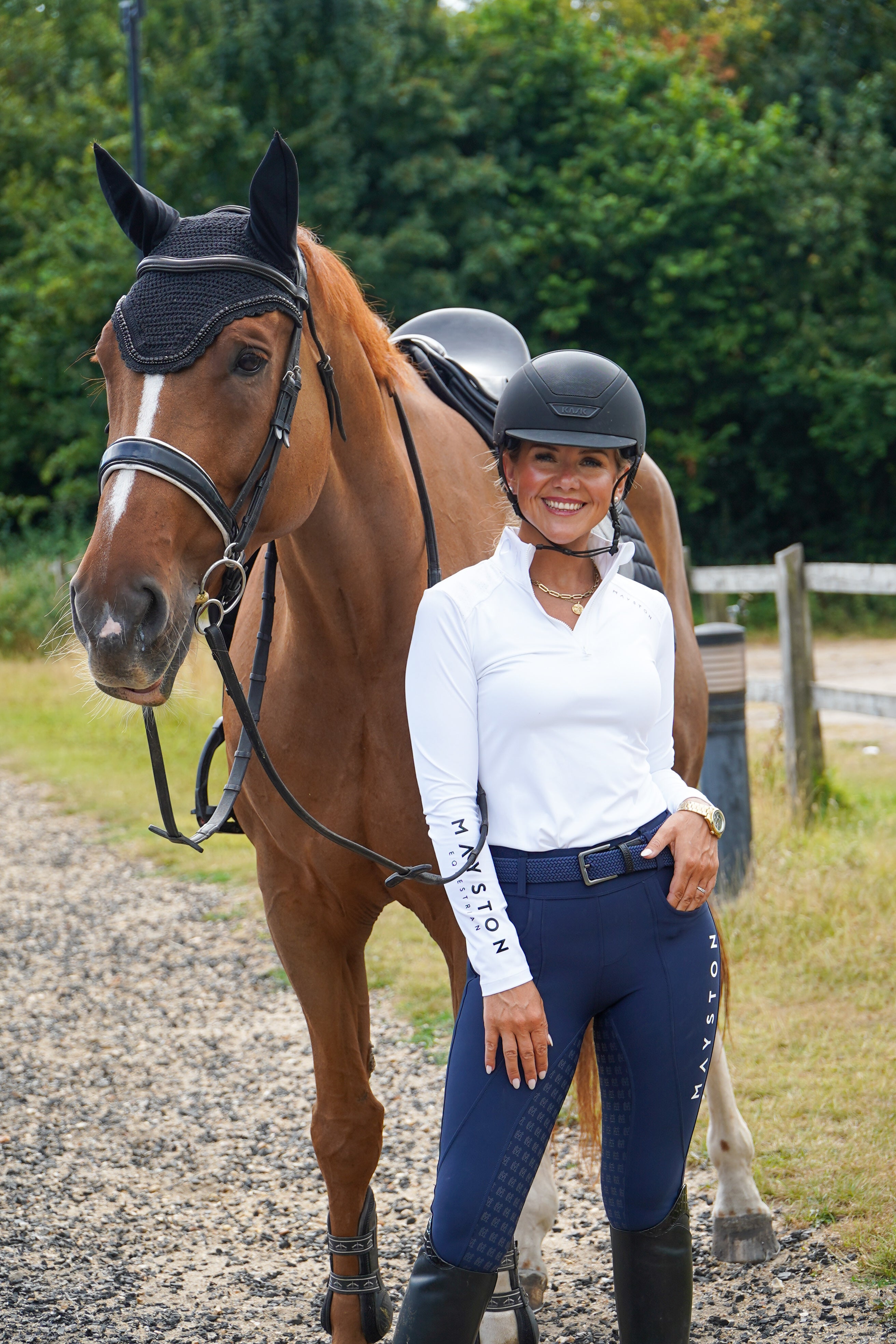 Woman in equestrian attire standing next to a brown horse on a gravel path with greenery in the background. Premium navy breeches with a Full Silicone seat. Stretch high waisted Jodhpurs. Deep pockets, supportive, comfortable and compression quality fabric. Elegant detailing with subtle branding. high gloss white branding