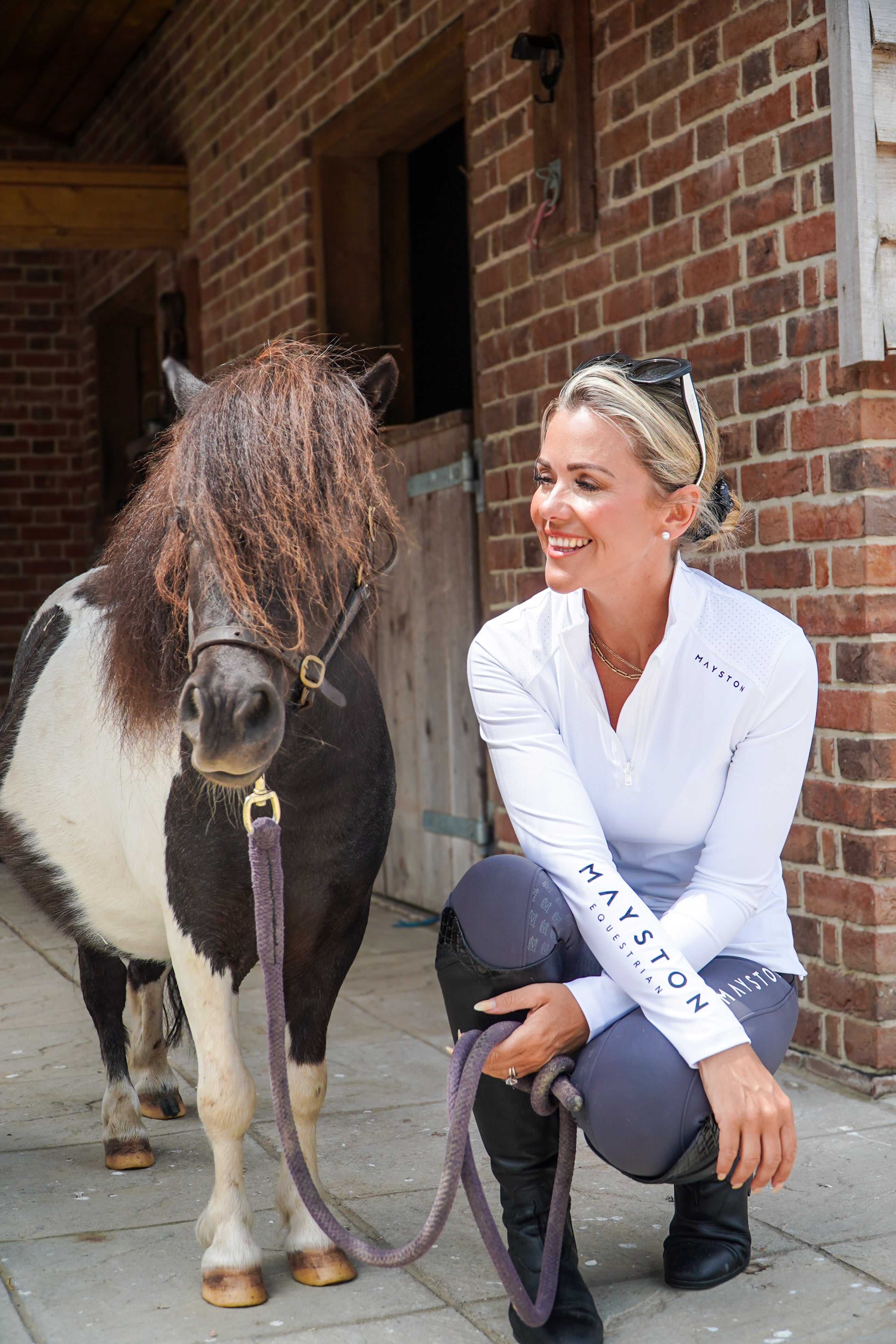 Woman in equestrian attire kneeling next to a small horse in front of a brick building. Sleek silhouette baselayer showshirt top subtle Mayston branding, maximum coverage without compromising style,  next-level performance with a bold, minimalist look. Moisture-wicking, breathable fabric with four-way stretch, it keeps you cool and comfortable. Training, Everyday or competition wear. long sleeve Sleeve 1/4 zip with standup collar. Suble Black Mayston branding.