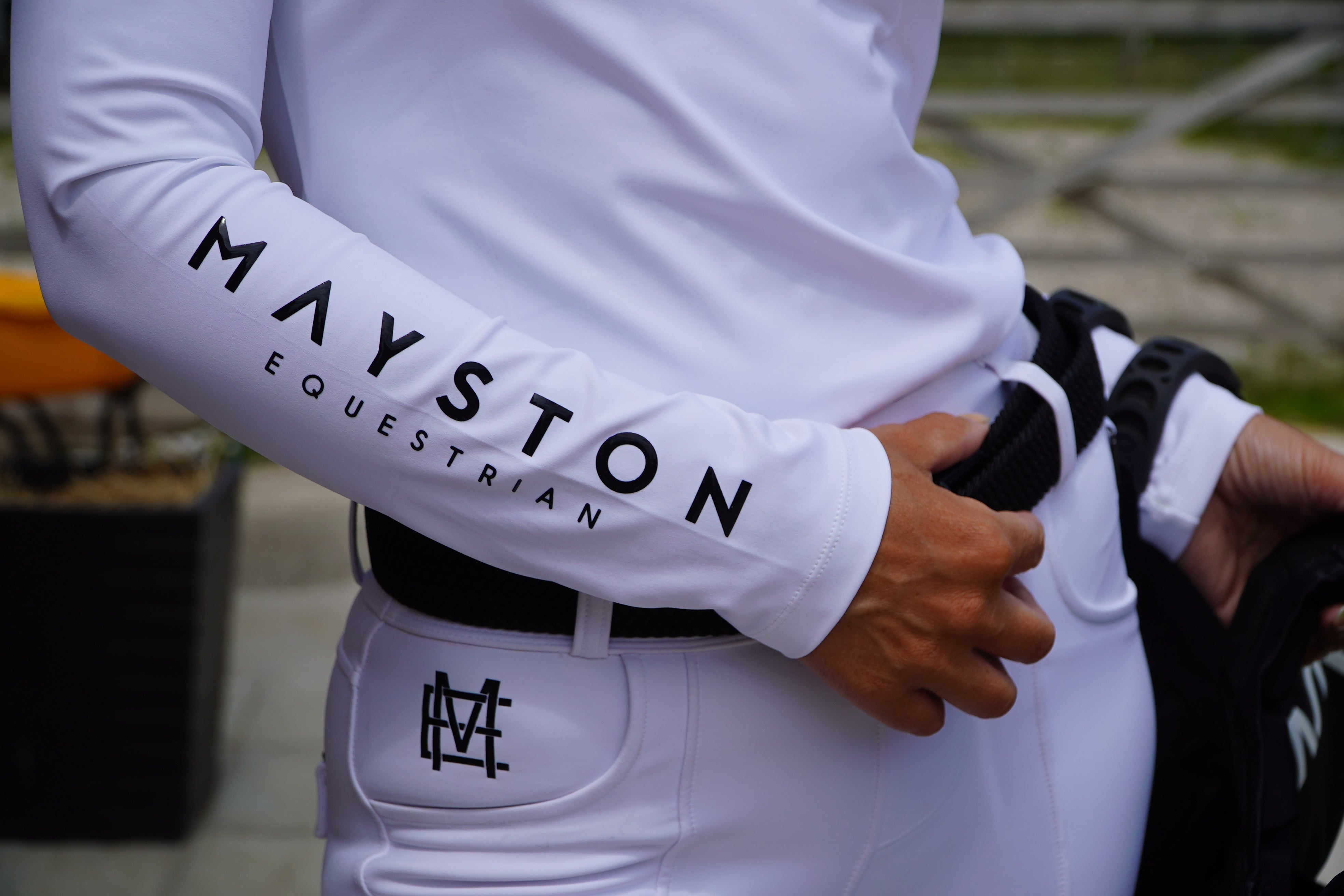 Person wearing a white equestrian outfit with 'Mayston Equestrian' branding. Sleek silhouette baselayer showshirt top subtle Mayston branding, maximum coverage without compromising style,  next-level performance with a bold, minimalist look. Moisture-wicking, breathable fabric with four-way stretch, it keeps you cool and comfortable. Training, Everyday or competition wear. long sleeve Sleeve 1/4 zip with standup collar. Suble Black Mayston branding.