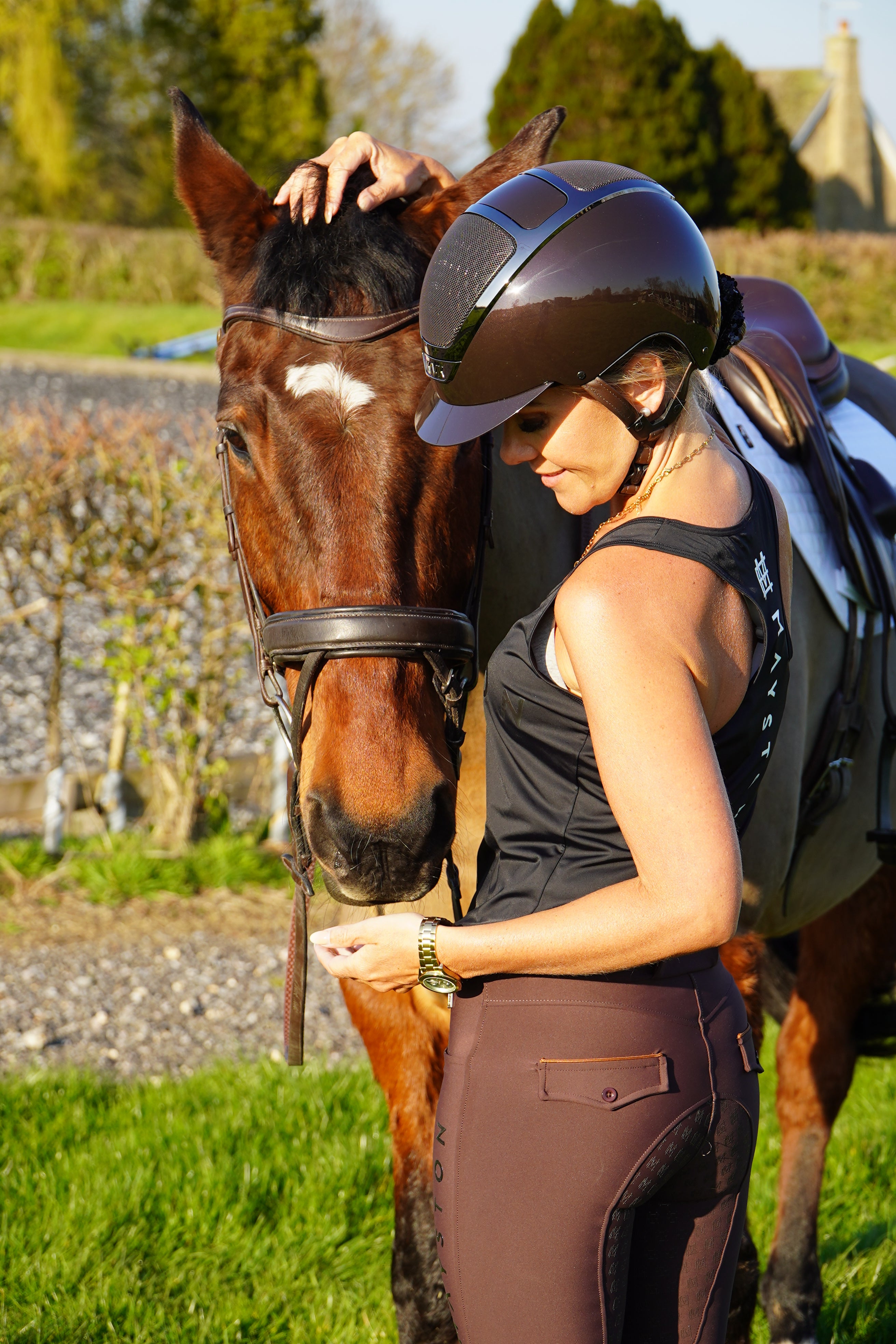 Woman in equestrian gear petting a horse by a stone wall and grass.  Radipole Racerback Vest, crafted from lightweight, high-quality sports material for effortless comfort and performance. Designed to move with you, it offers breathability and freedom of motion, making it perfect for riding, training, or everyday wear. Finished with bold white high-gloss Mayston silicone branding on the chest and a sleek logo at the rear, this black vest combines sporty style with signature Mayston.