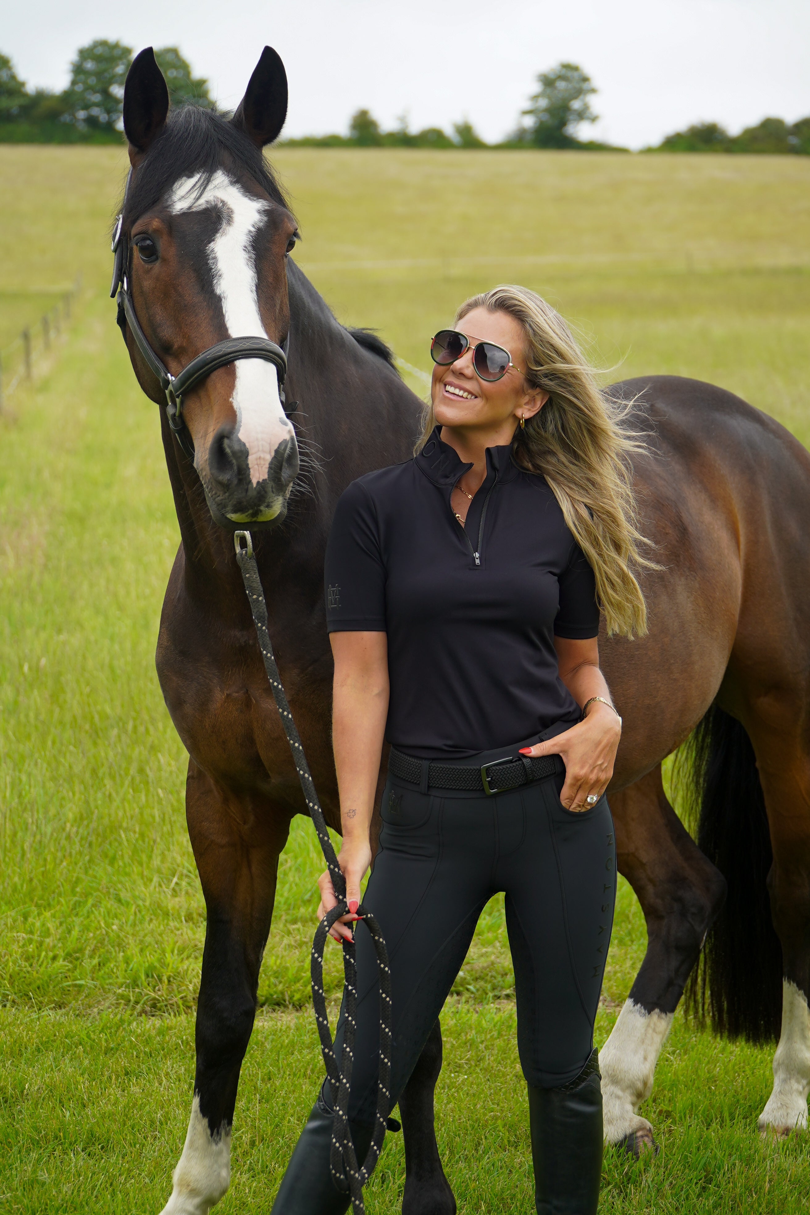 Woman standing next to a horse in a grassy field. Broadstone Short sleeve Luxury Sports material base layer short sleeve. Black with subtle black branding colour match embroidery. High collar that's not restrictive. 1/4 zip. Blackout design