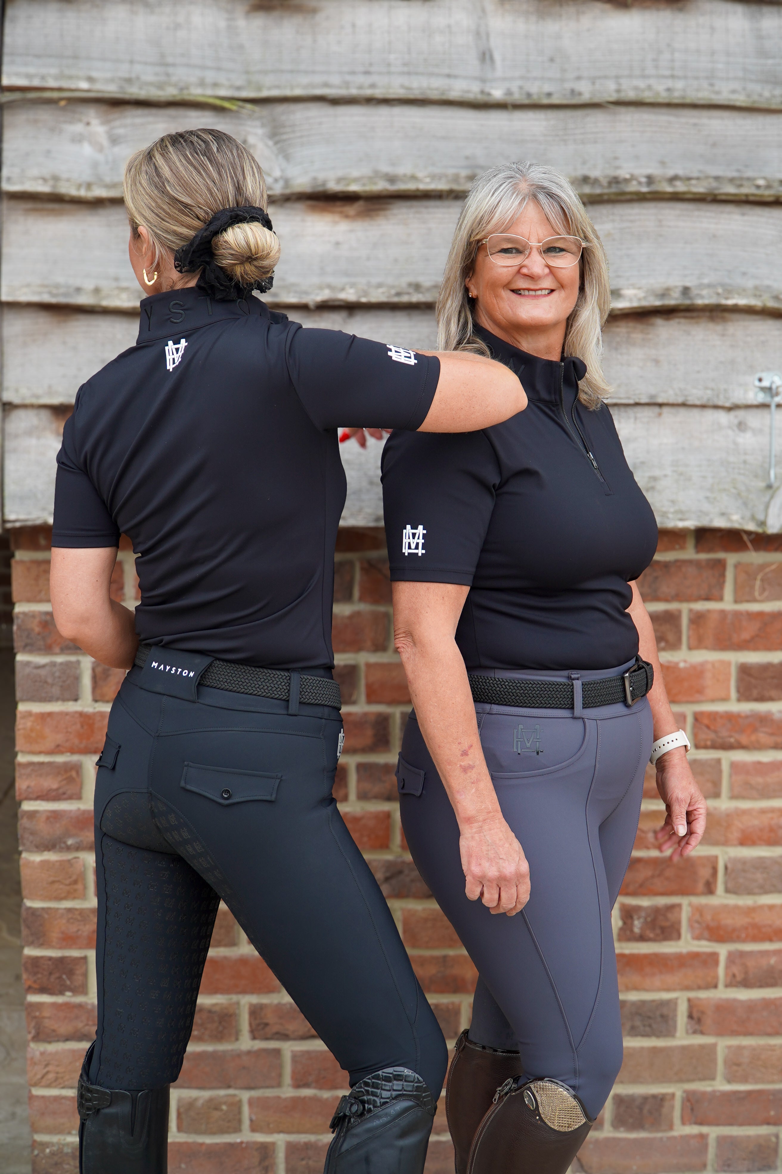 Two women in equestrian attire standing against a brick wall. Broadstone Short sleeve Luxury Sports material base layer short sleeve. Black with subtle white branding colour match embroidery. High collar that's not restrictive. 1/4 zip design
