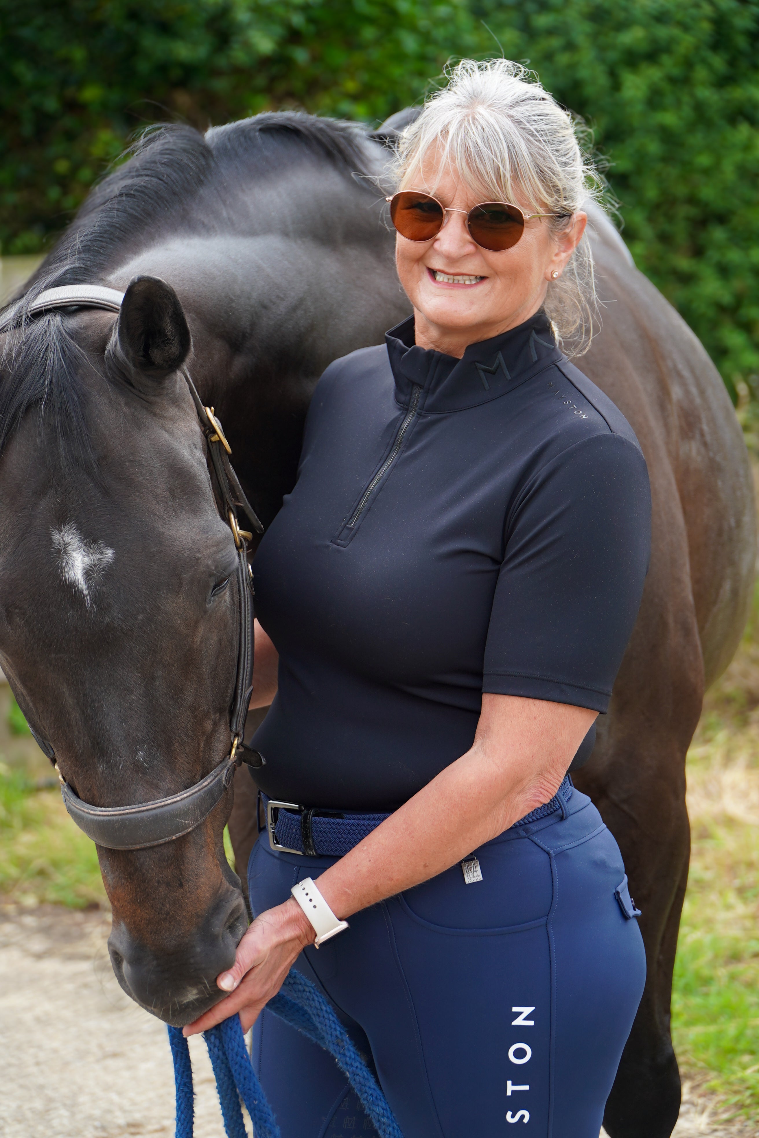 Woman in equestrian attire standing next to a horse outdoors. Broadstone Short sleeve Luxury Sports material base layer short sleeve. Black with subtle black branding colour match embroidery. High collar that's not restrictive. 1/4 zip. Blackout design