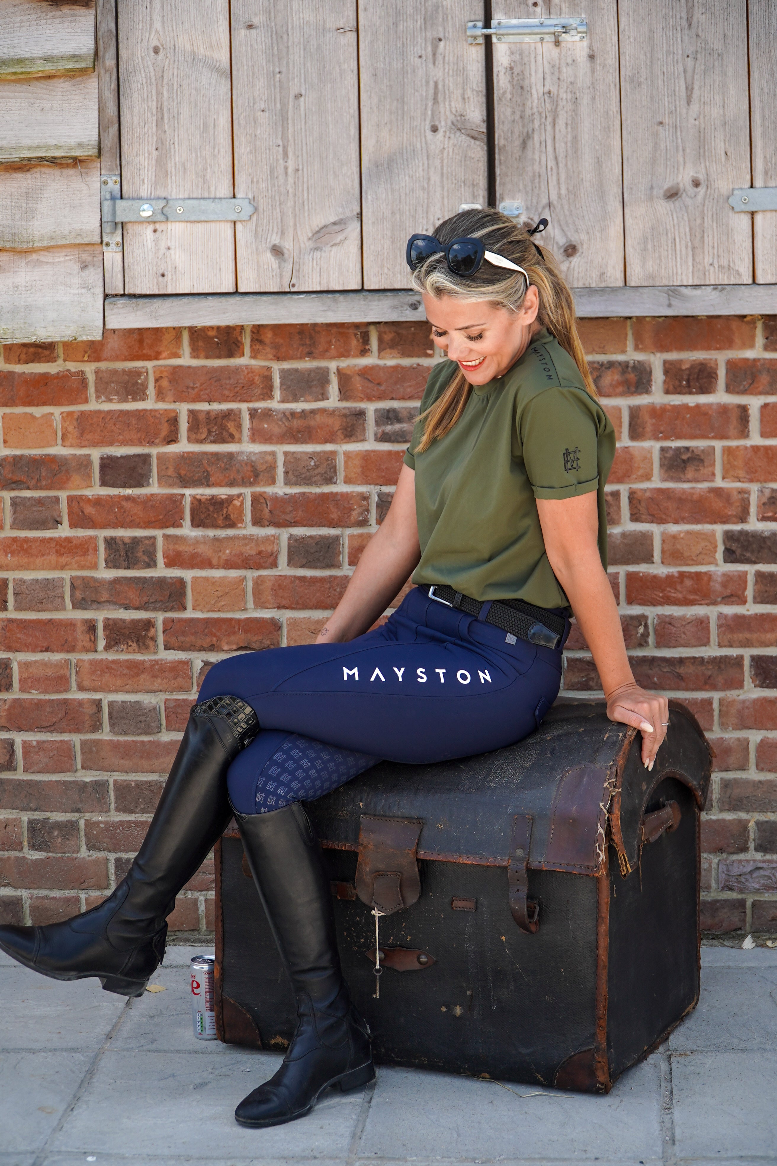 Woman in equestrian attire sitting on a suitcase against a brick wall. luxury quality t shirt and fabric is specially crafted for active duty, providing flexibility and ease of movement for any busy lifestyle. An ideal choice for a day at the yard, in the saddle, or during your grooming duties, its loose-fitting design guarantees complete freedom. Hunter green colour