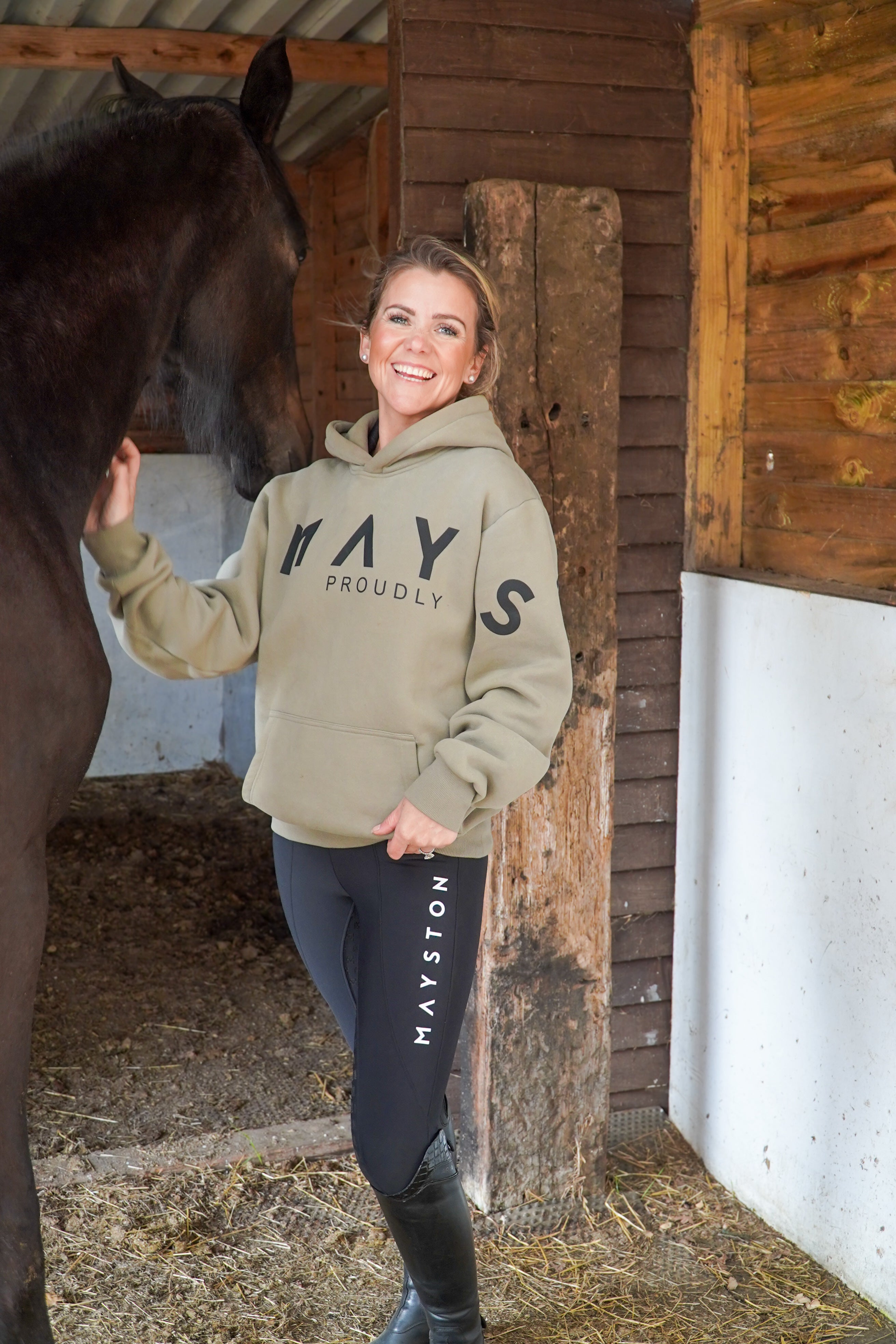 Woman in a green hoodie and black breeches standing next to a horse in a stable. High quality luxurious 100% cotton. wrap around bold logo. soft, cozy and  warm. Hoodie, jumper sweatshirt. everyday wear. casual and smart