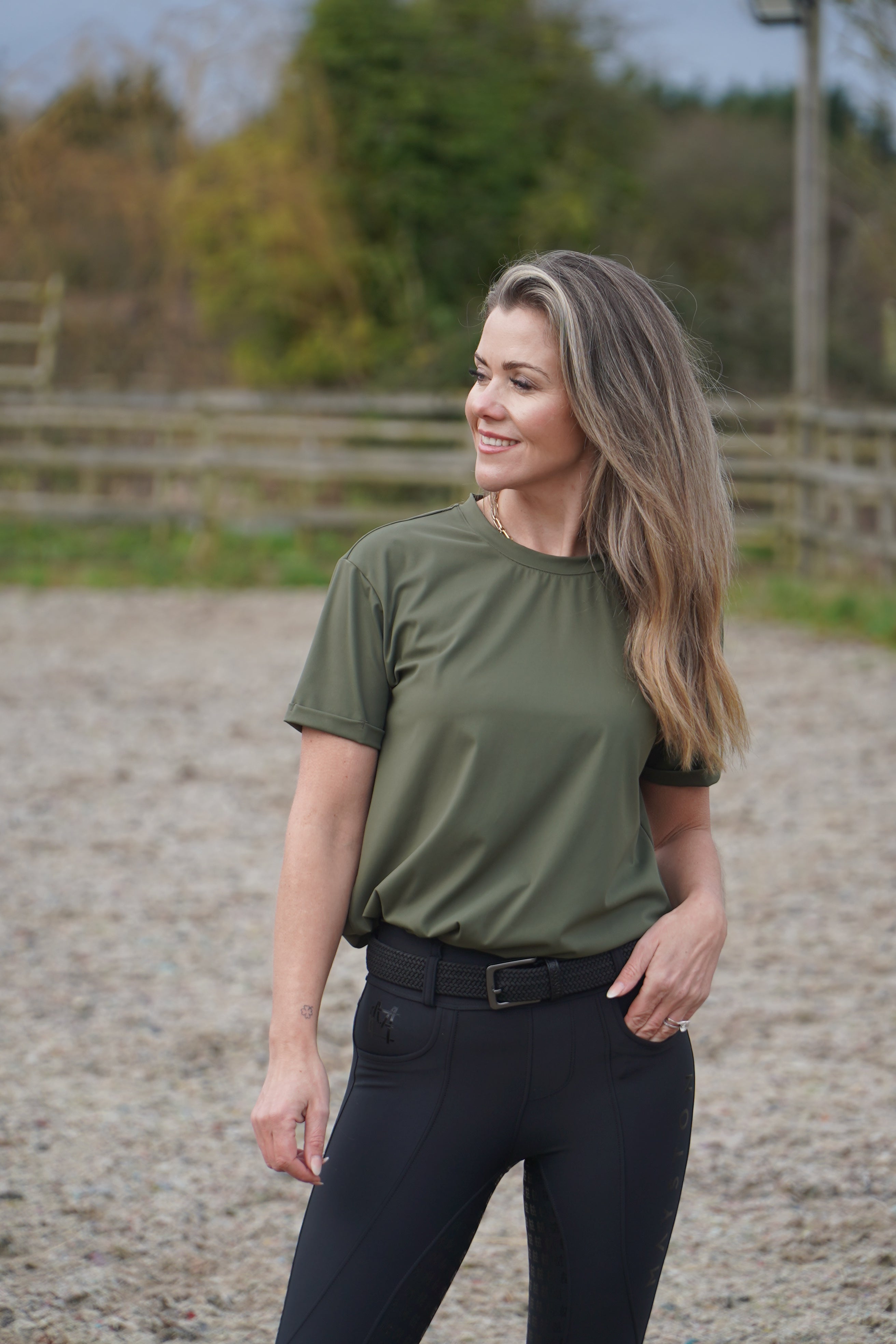 Woman in green shirt and black pants standing outdoors with a blurred background. luxury quality t shirt and fabric is specially crafted for active duty, providing flexibility and ease of movement for any busy lifestyle. An ideal choice for a day at the yard, in the saddle, or during your grooming duties, its loose-fitting design guarantees complete freedom. Hunter green colour