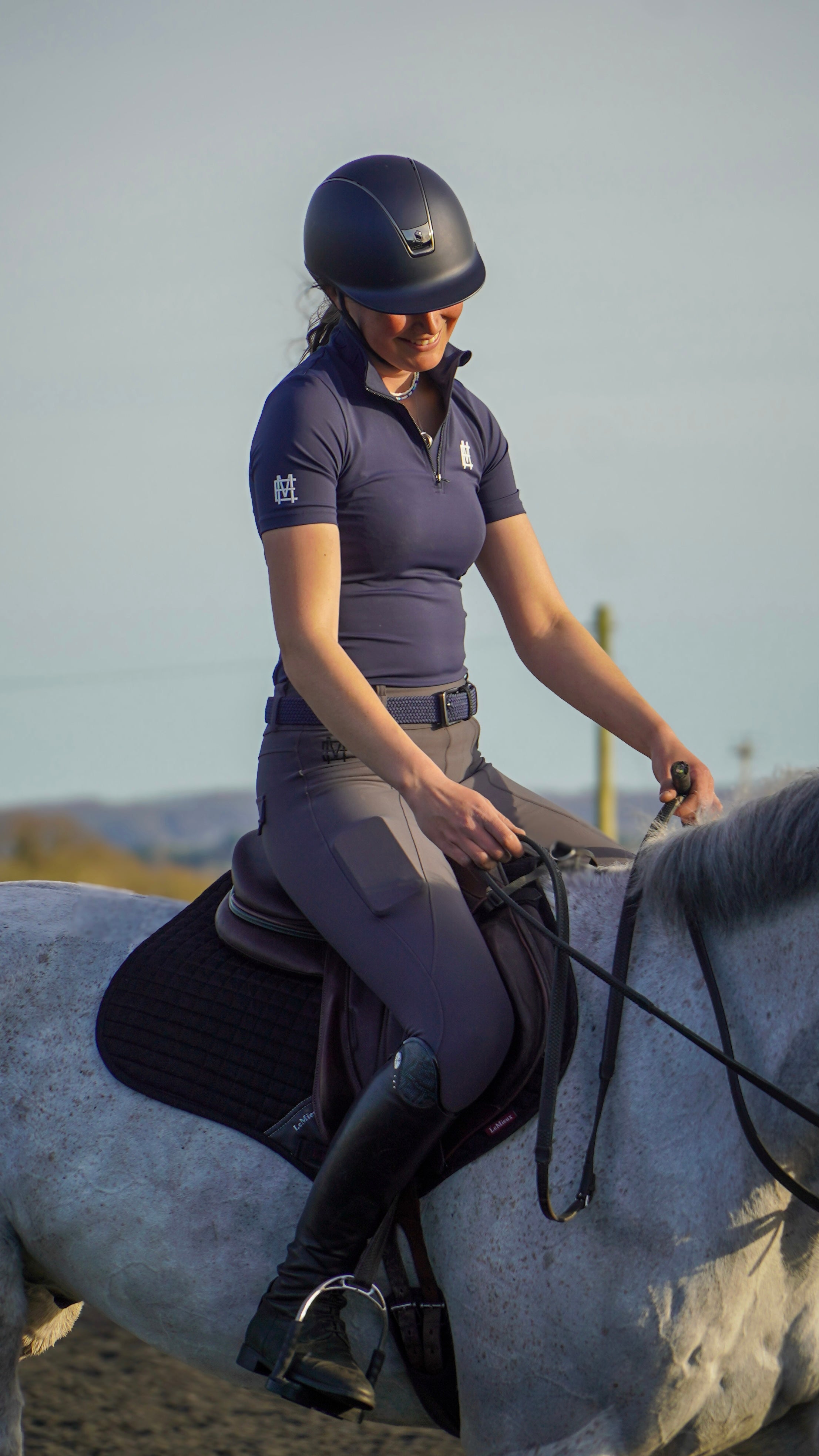 Sleek silhouette baselayer top subtle Mayston branding, maximum coverage without compromising style,  next-level performance with a bold, minimalist look. Moisture-wicking, breathable fabric with four-way stretch, it keeps you cool and comfortable. Training, Everyday or competition wear. Short Sleeve 1/4 zip with collar. navy with white branding. Woman riding grey horse