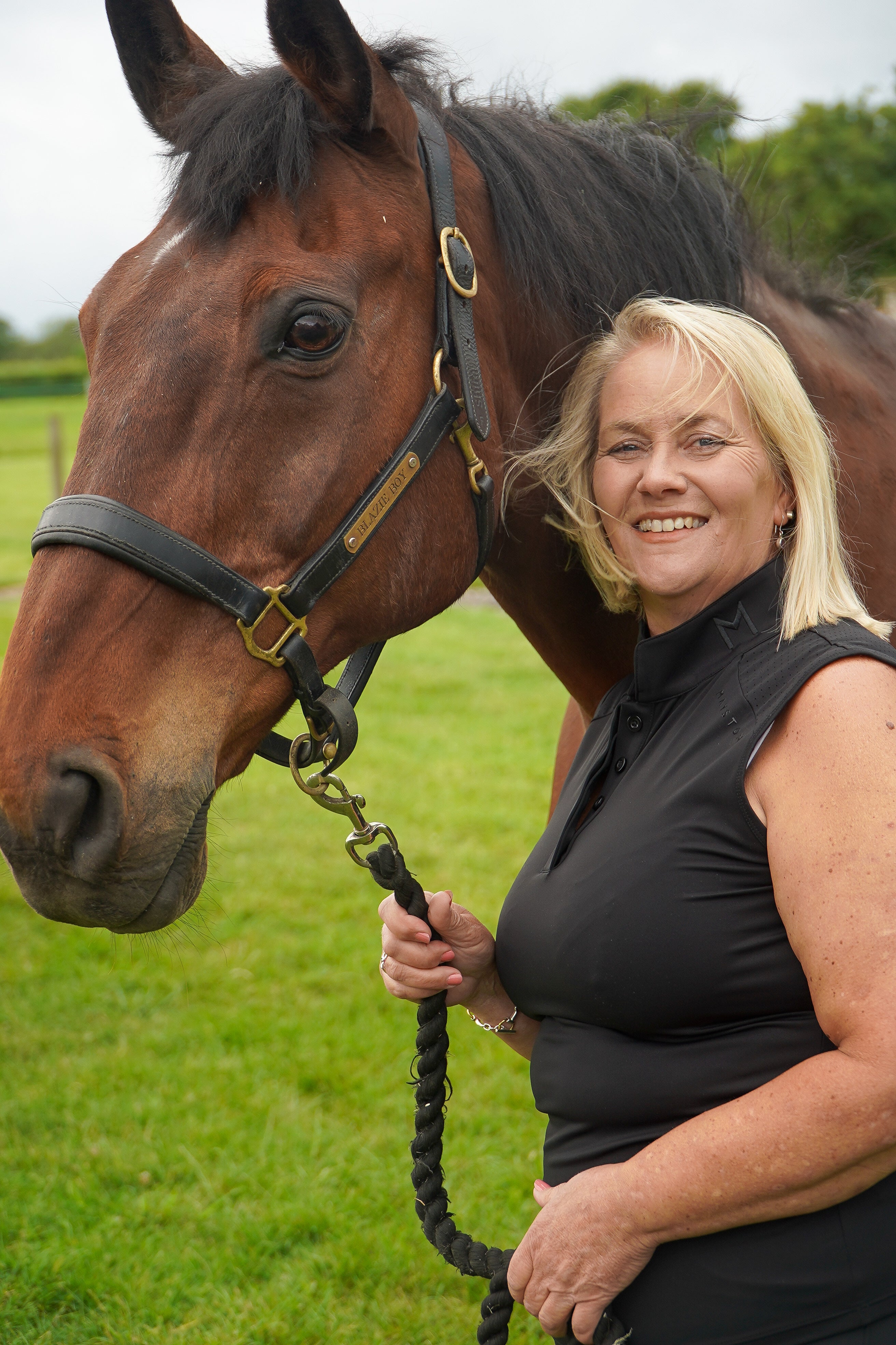 Woman standing next to a horse in an outdoor setting.  Ultra-soft technical sport fabric. Breathable and lightweight for warmer weather. Flattering athletic fit
Signature black branded Mayston collar. Subtle gloss shoulder logo detail. Sleeveless design for unrestricted movement. blackout colour match branding