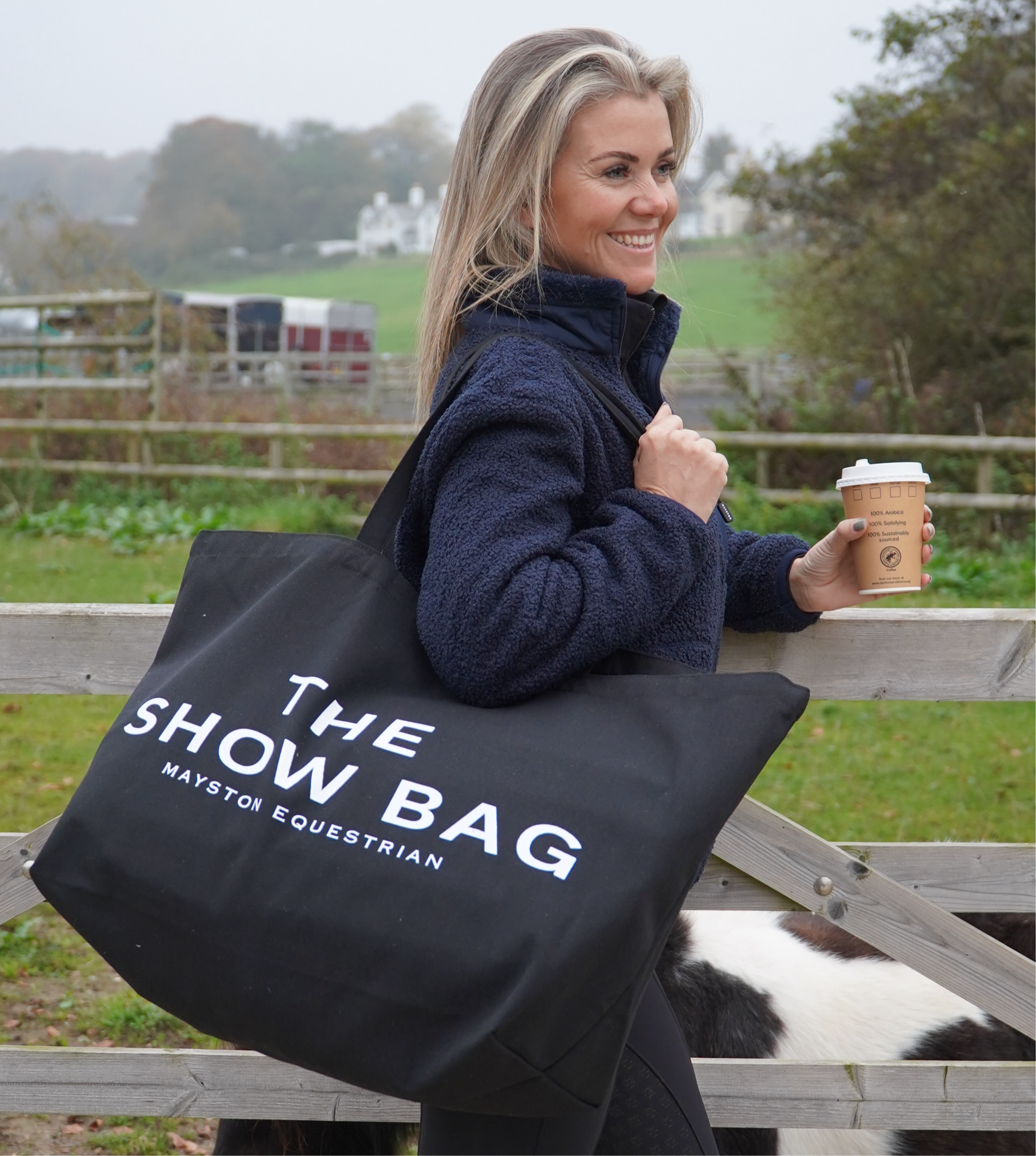 A woman carrying a black tote bag with the text 'THE SHOW BAG' printed on it, in a equestrian setting.