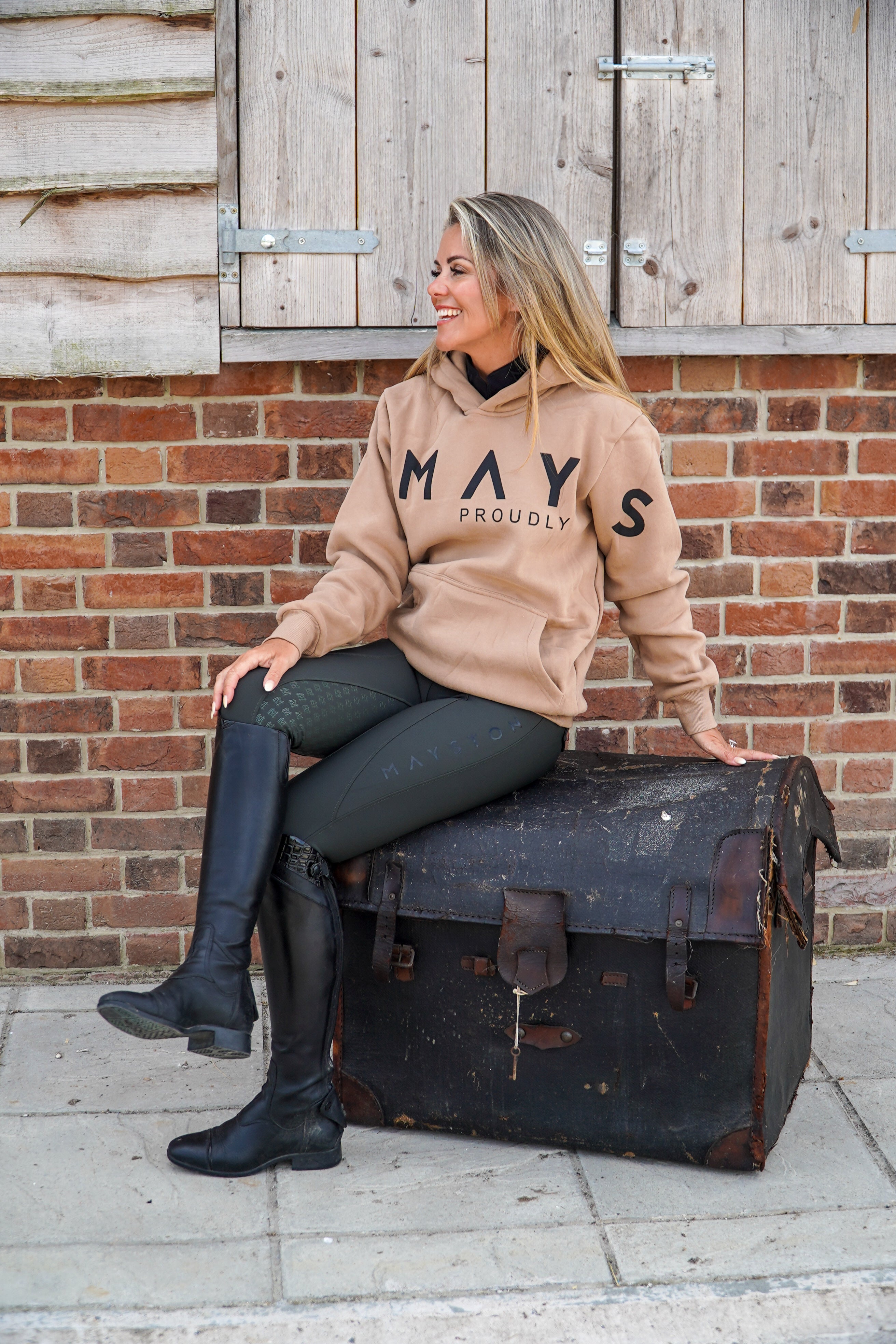 Woman sitting on a vintage suitcase wearing a beige hoodie with 'MAYSTON' branding against a brick wall. 
High quality luxurious 100% cotton. wrap around bold logo. soft, cozy and  warm. Hoodie, jumper sweatshirt. everyday wear. casual and smart