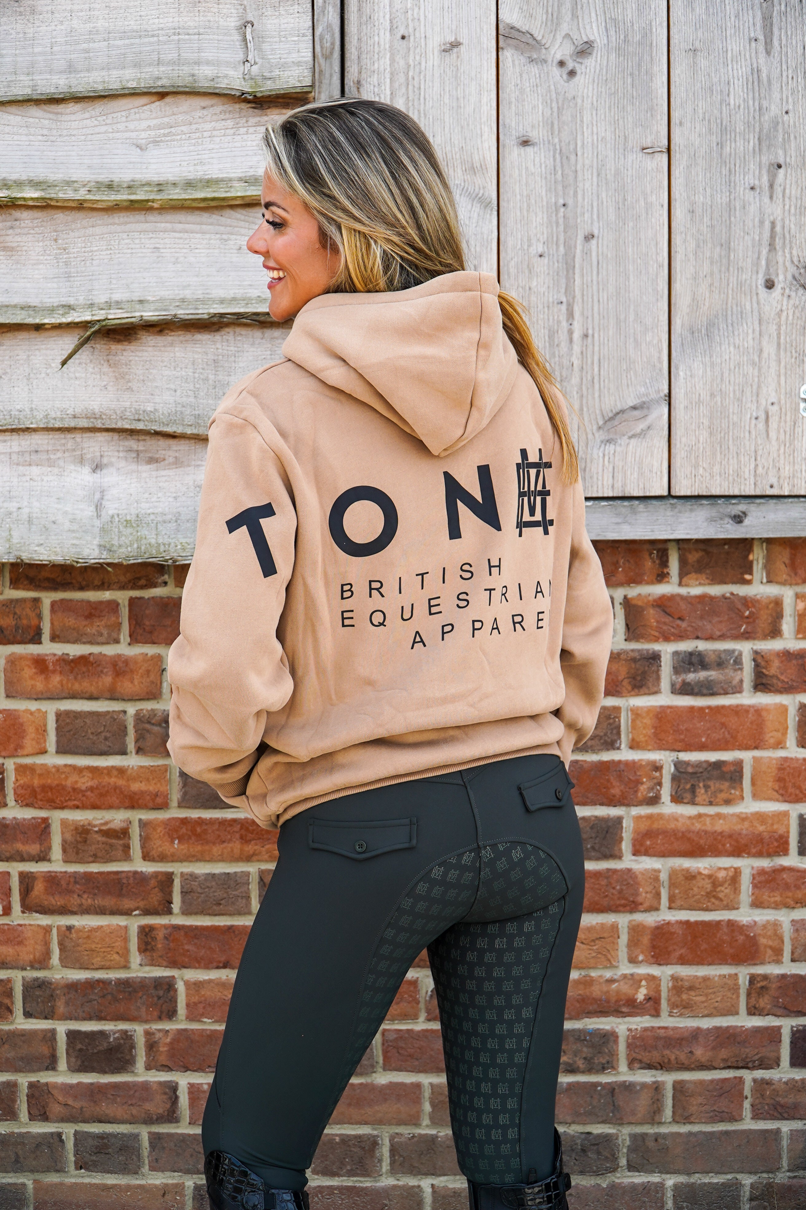 Woman wearing a beige hoodie with 'Tone British Equestrian Apparel' text, standing against a brick wall. High quality luxurious 100% cotton. wrap around bold logo. soft, cozy and  warm. Hoodie, jumper sweatshirt. everyday wear. casual and smart. brown and black and beige