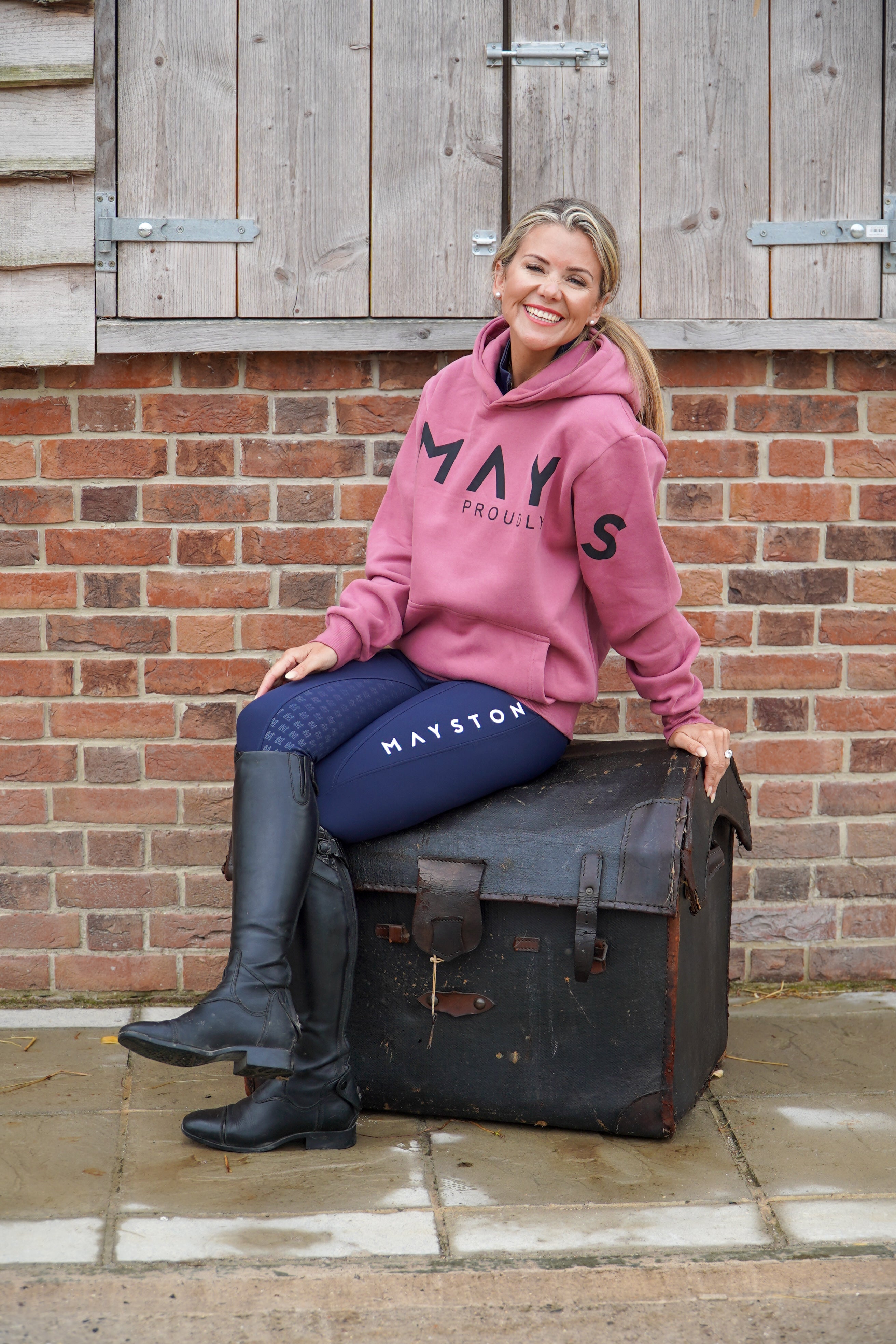 Woman in pink hoodie and blue breeches sitting on a suitcase with 'Mays' branding against a brick wall.
High quality luxurious 100% cotton. wrap around bold logo. soft, cozy and  warm. Hoodie, jumper sweatshirt. everyday wear. casual and smart