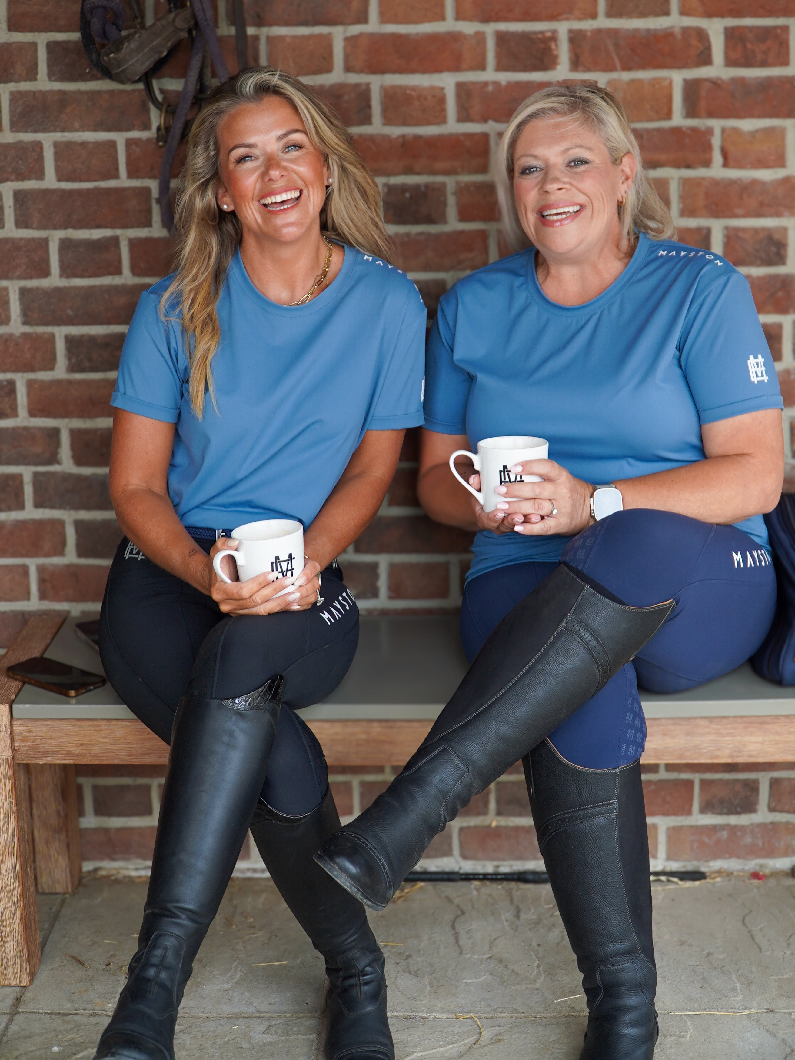 Two women in blue shirts and riding pants sitting on a bench with mugs, against a brick wall. luxury quality t shirt and fabric is specially crafted for active duty, providing flexibility and ease of movement for any busy lifestyle. An ideal choice for a day at the yard, in the saddle, or during your grooming duties, its loose-fitting design guarantees complete freedom.