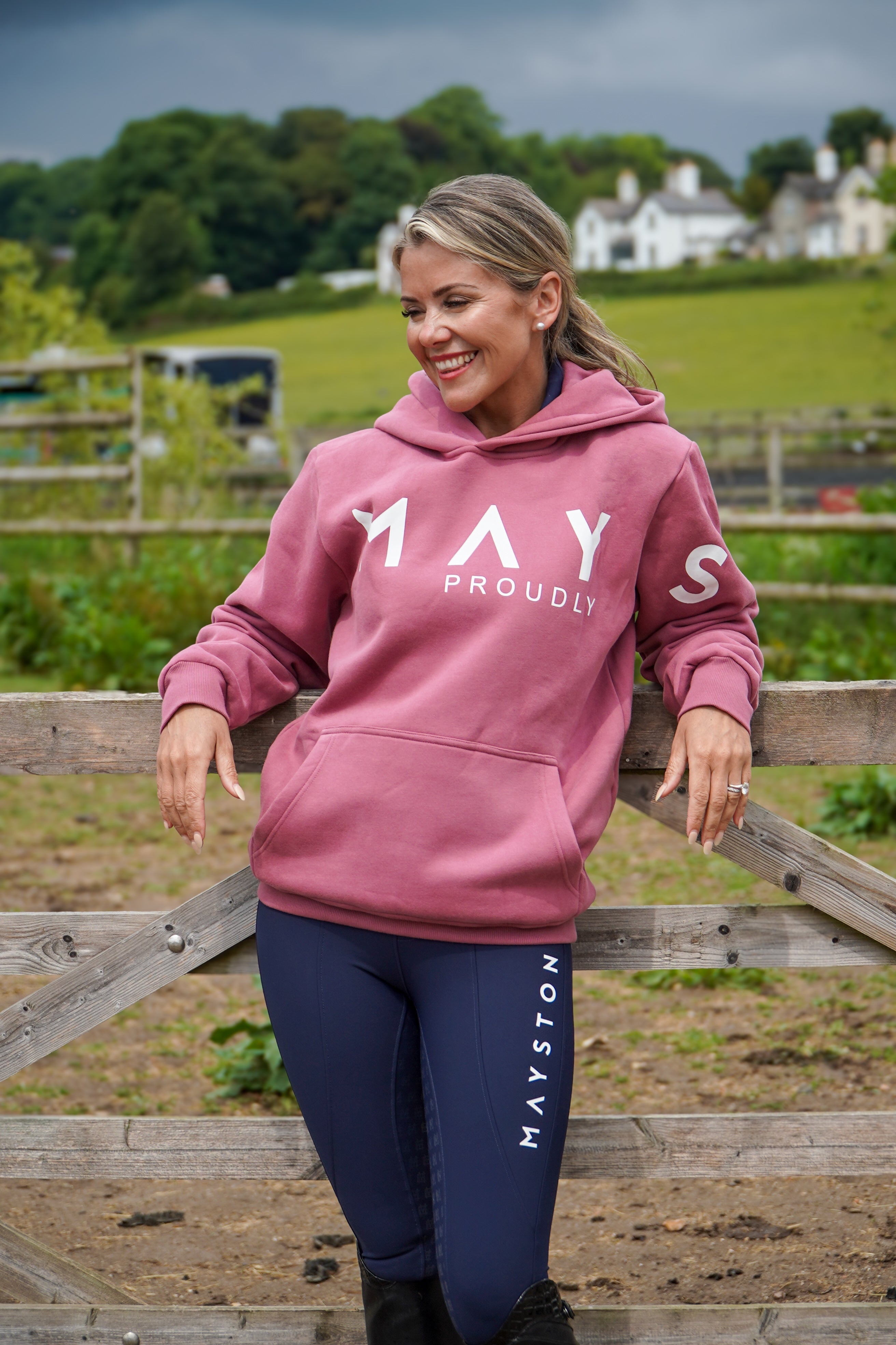 Woman wearing a pink hoodie with 'MAY PROUDLY' text and blue breeches with 'MAVSTON' text, standing in a field. High quality luxurious 100% cotton. wrap around bold logo. soft, cozy and  warm. Hoodie, jumper sweatshirt. everyday wear. casual and smart