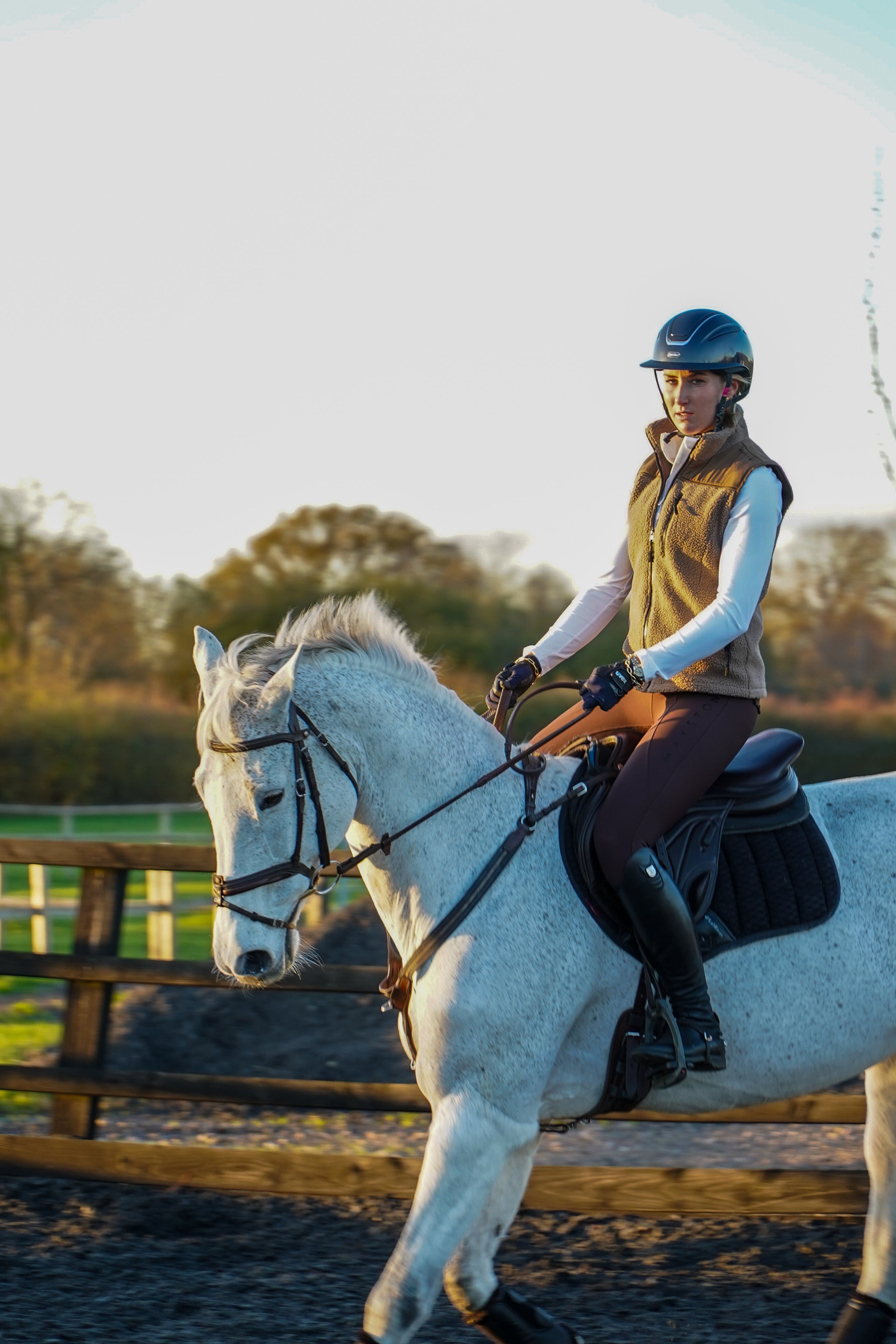 Person riding a white horse in an outdoor setting with trees and a fence in the background. Luxury teddy Fleece Gilet Waistcoat. Quality embroidery. multiple pockets. Subtle branding, elegant classic design. Stretch lining