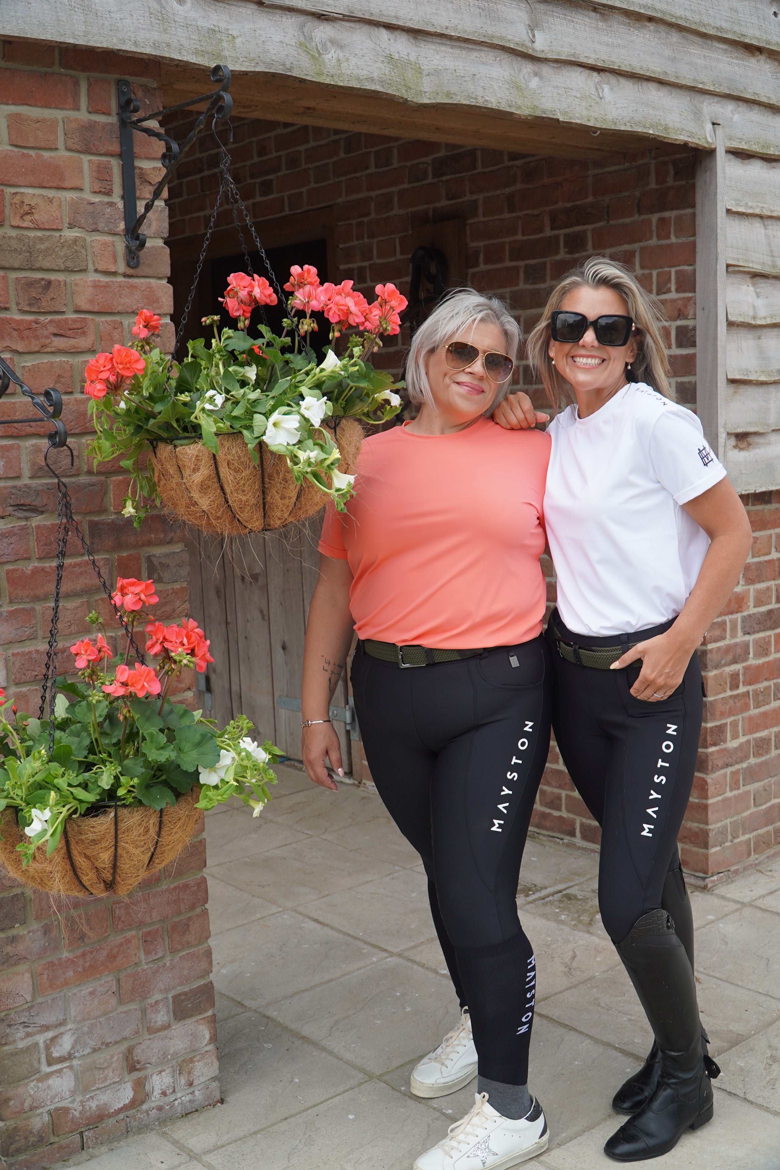 Two women standing on a porch with plants and a door in the background. luxury white and pink quality t shirt and fabric is specially crafted for active duty, providing flexibility and ease of movement for any busy lifestyle. An ideal choice for a day at the yard, in the saddle, or during your grooming duties, its loose-fitting design guarantees complete freedom.