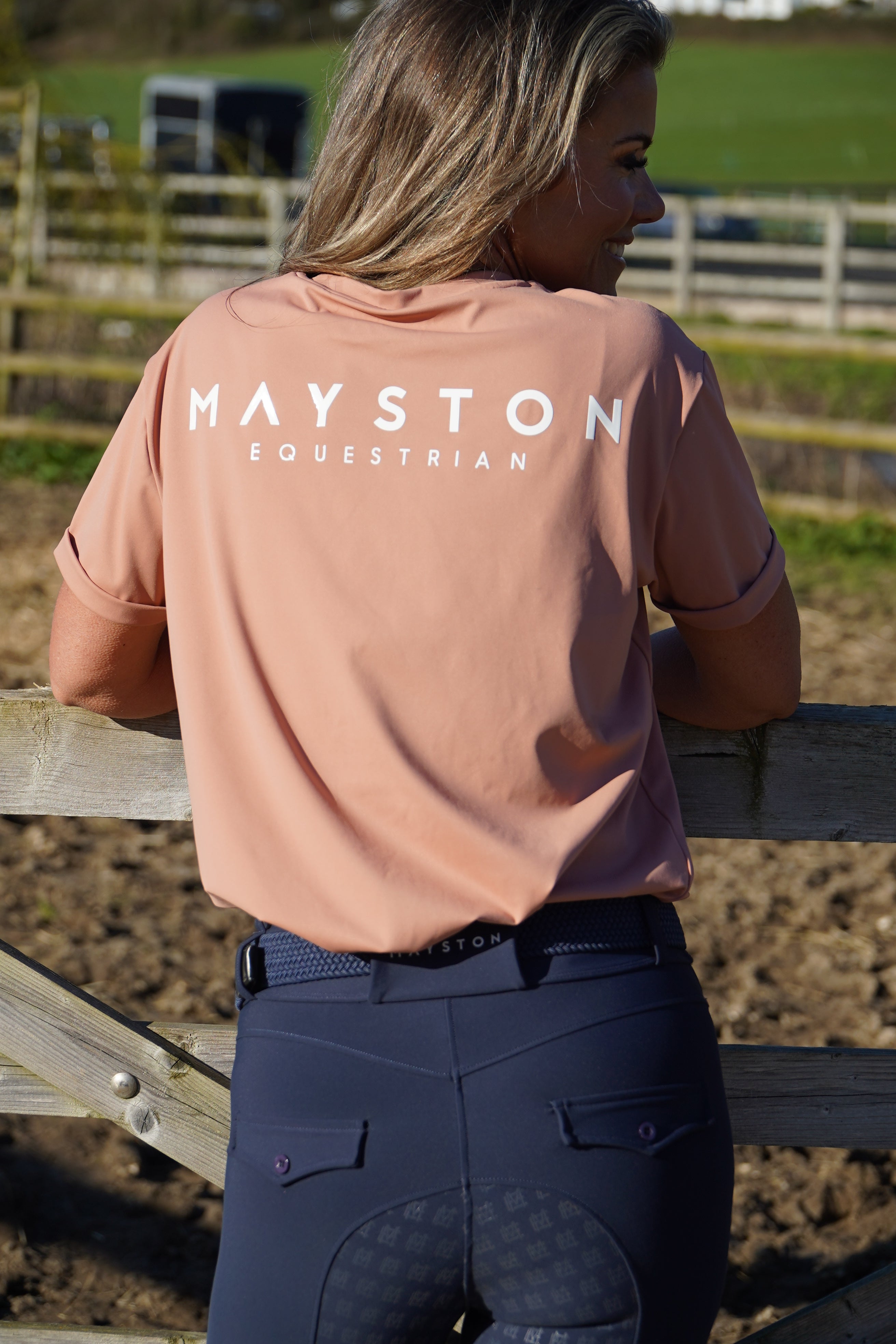 Person wearing a pink Mayston Equestrian jacket outdoors. luxury quality pink blush t shirt and fabric is specially crafted for active duty, providing flexibility and ease of movement for any busy lifestyle. An ideal choice for a day at the yard, in the saddle, or during your grooming duties, its loose-fitting design guarantees complete freedom. high gloss silicone branding 