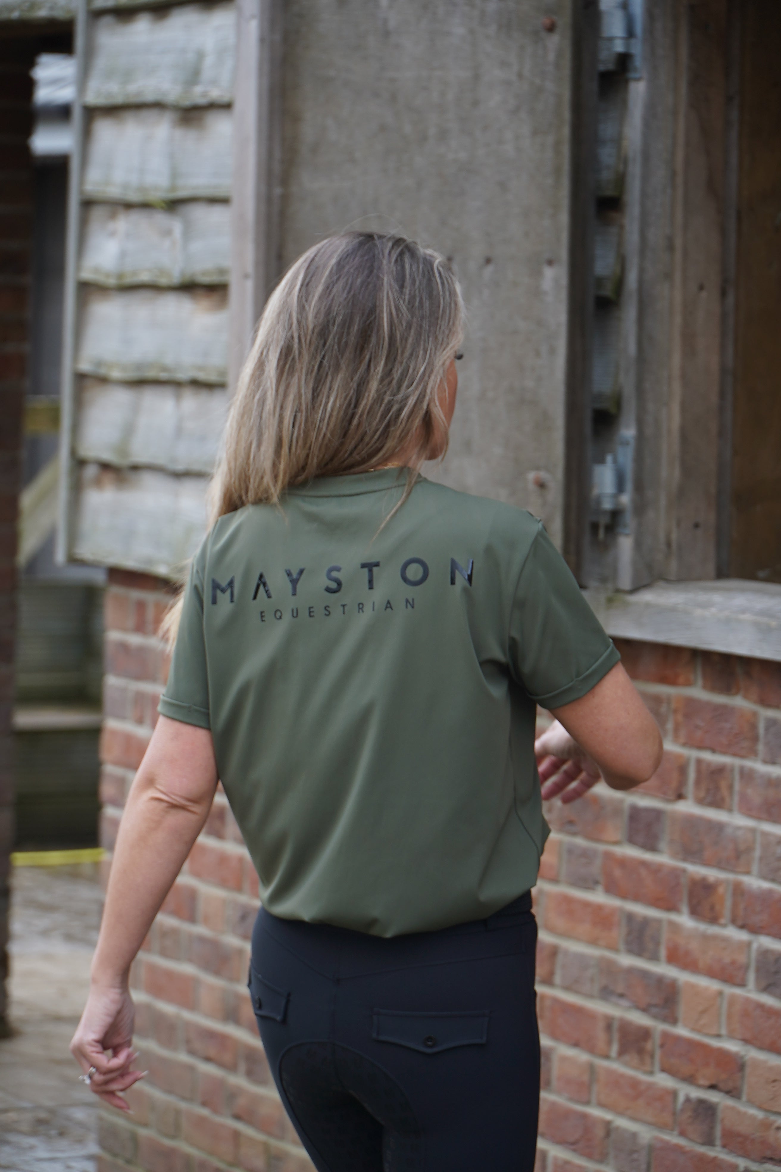Person wearing a green shirt with 'Mayston' branding, standing in front of a brick wall. luxury quality t shirt and fabric is specially crafted for active duty, providing flexibility and ease of movement for any busy lifestyle. An ideal choice for a day at the yard, in the saddle, or during your grooming duties, its loose-fitting design guarantees complete freedom. Hunter green colour